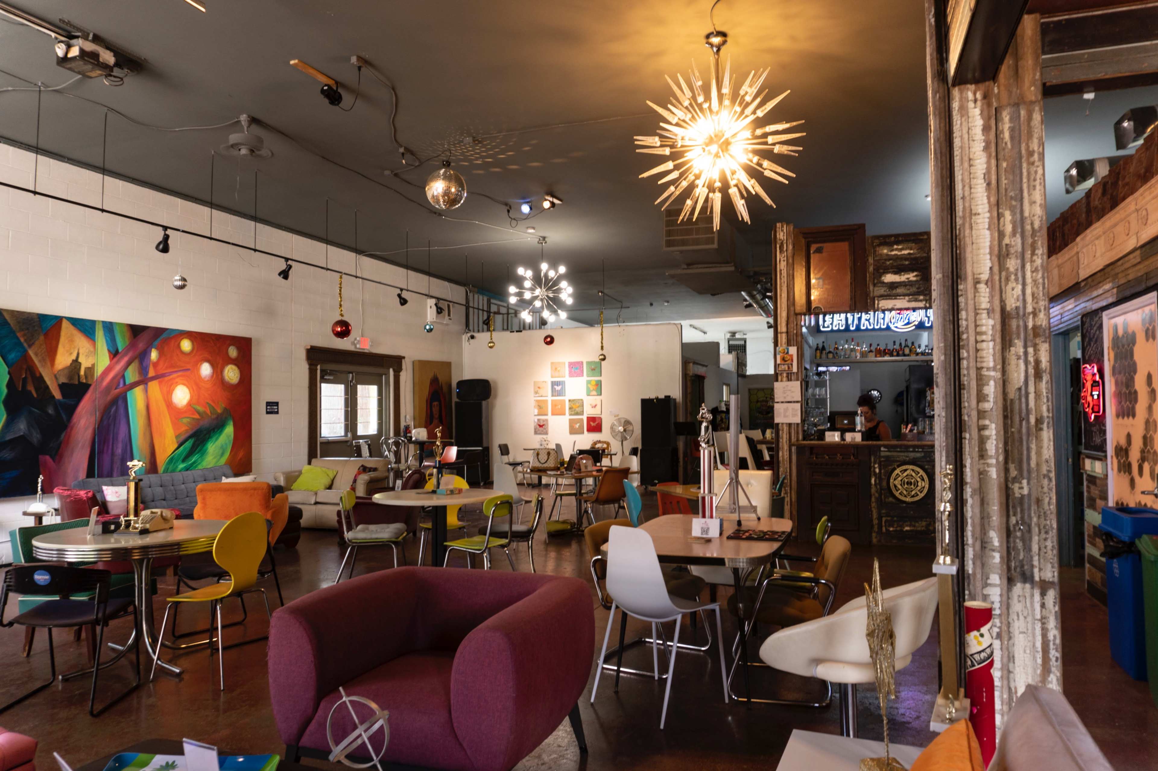 The image shows a vibrant café interior featuring colorful furniture, artistic wall decor, and unique lighting fixtures.