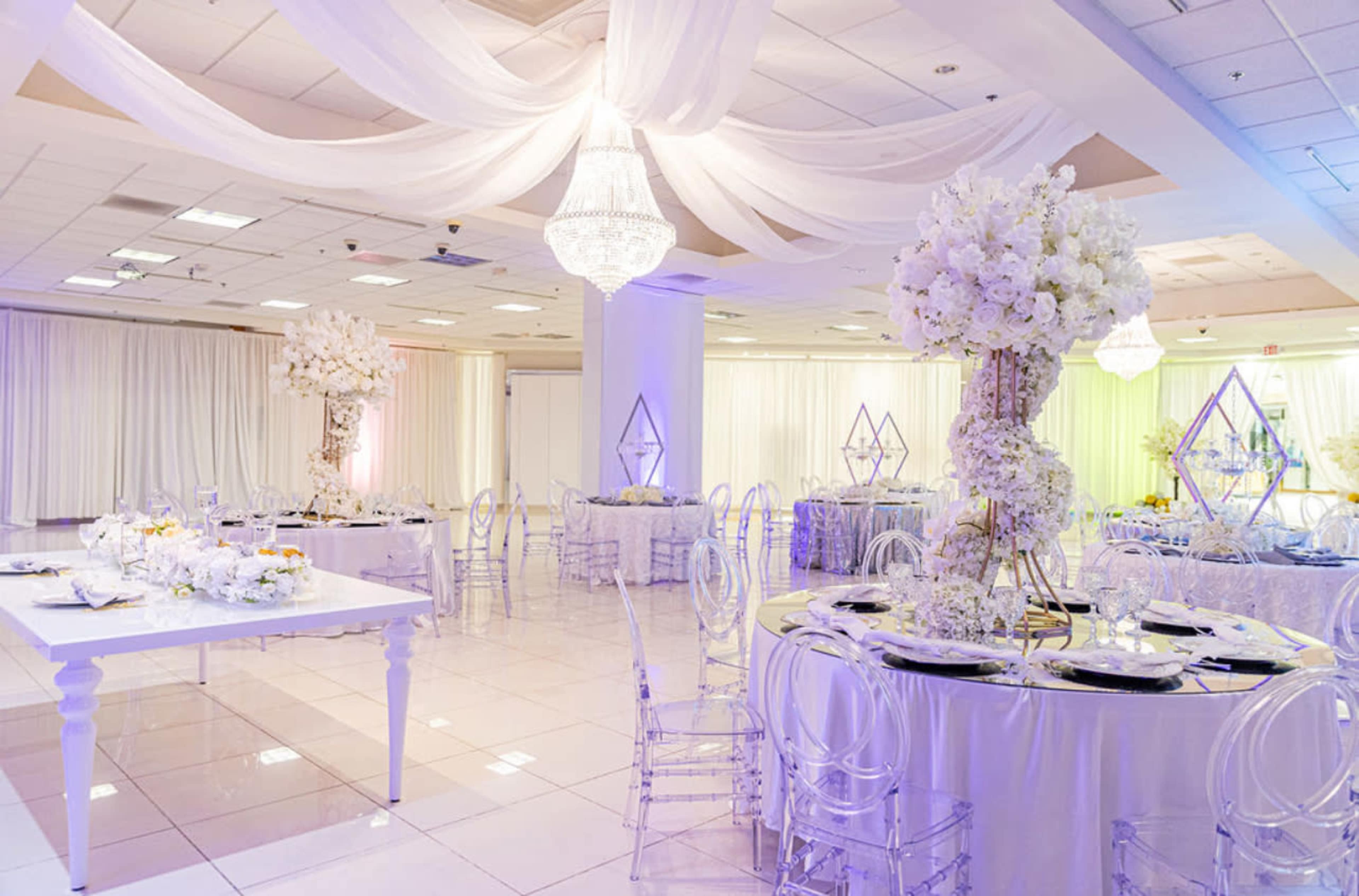 An elegantly decorated banquet hall with white table settings, floral centerpieces, and draped fabric overhead.