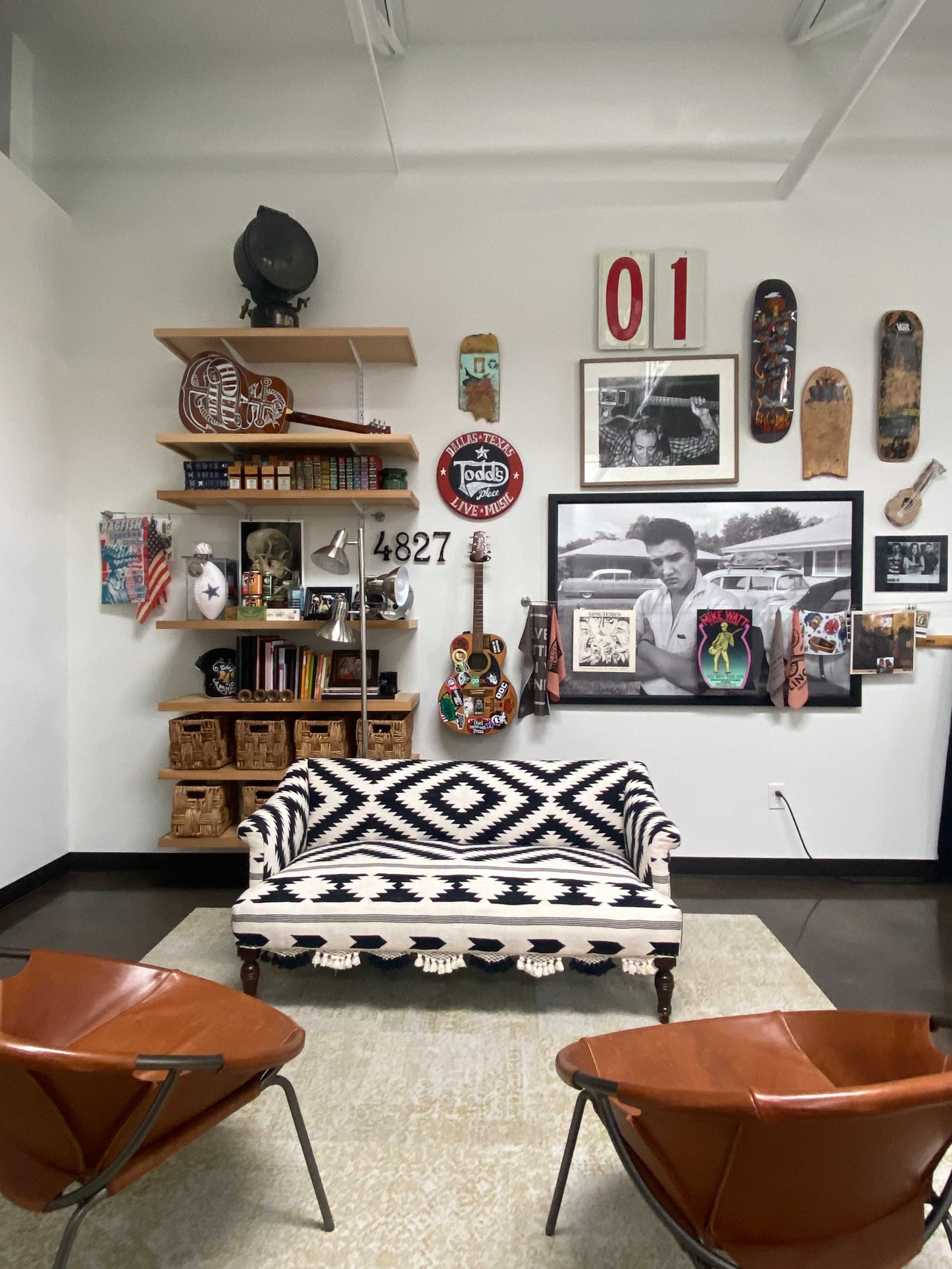 A modern living room with a patterned couch, two leather chairs, and a decorated wall featuring shelves, artwork, and a guitar.