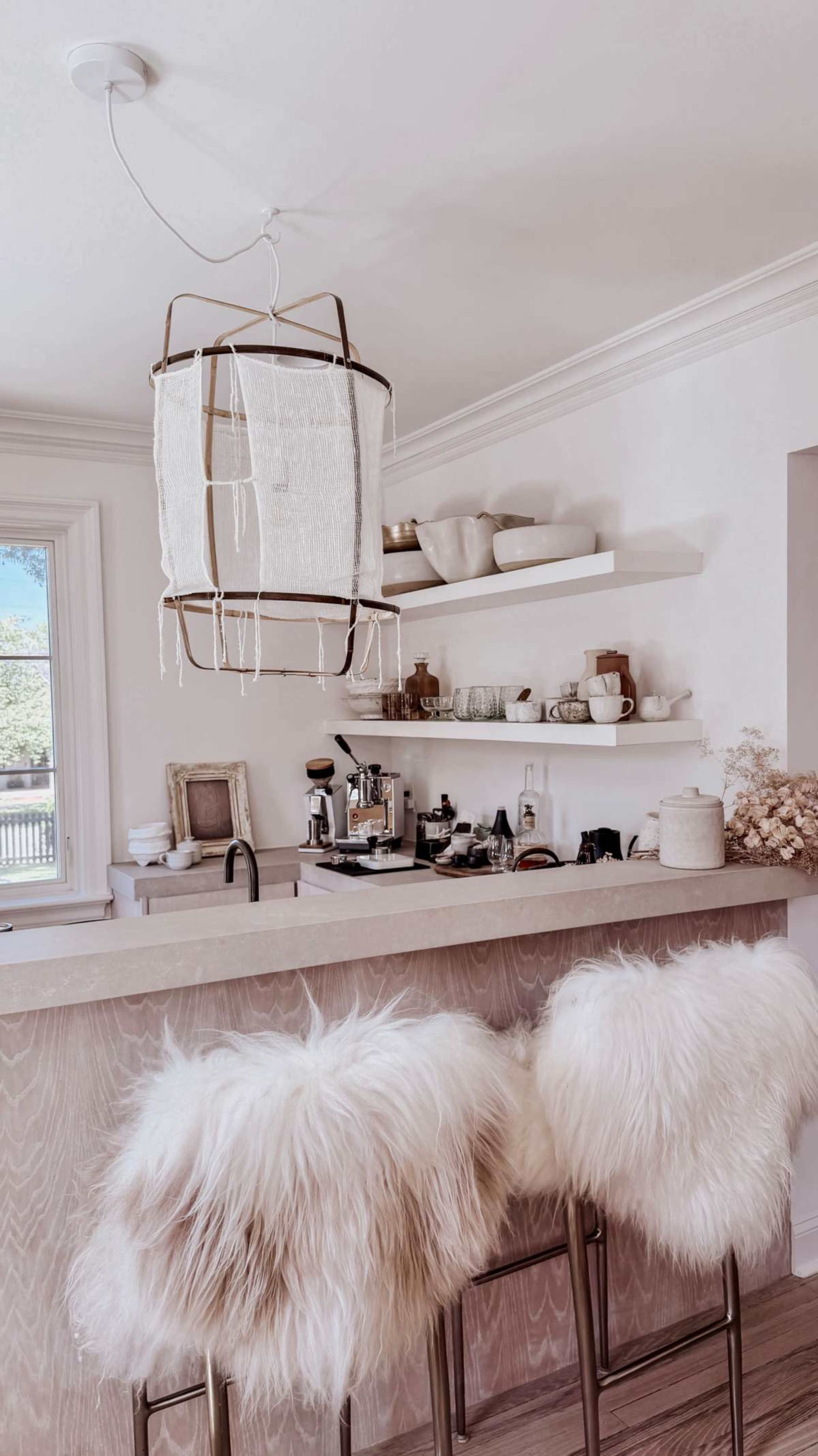 A modern kitchen features a light fixture above a counter with two bar stools adorned with fur, while shelves display dishware and kitchen appliances.