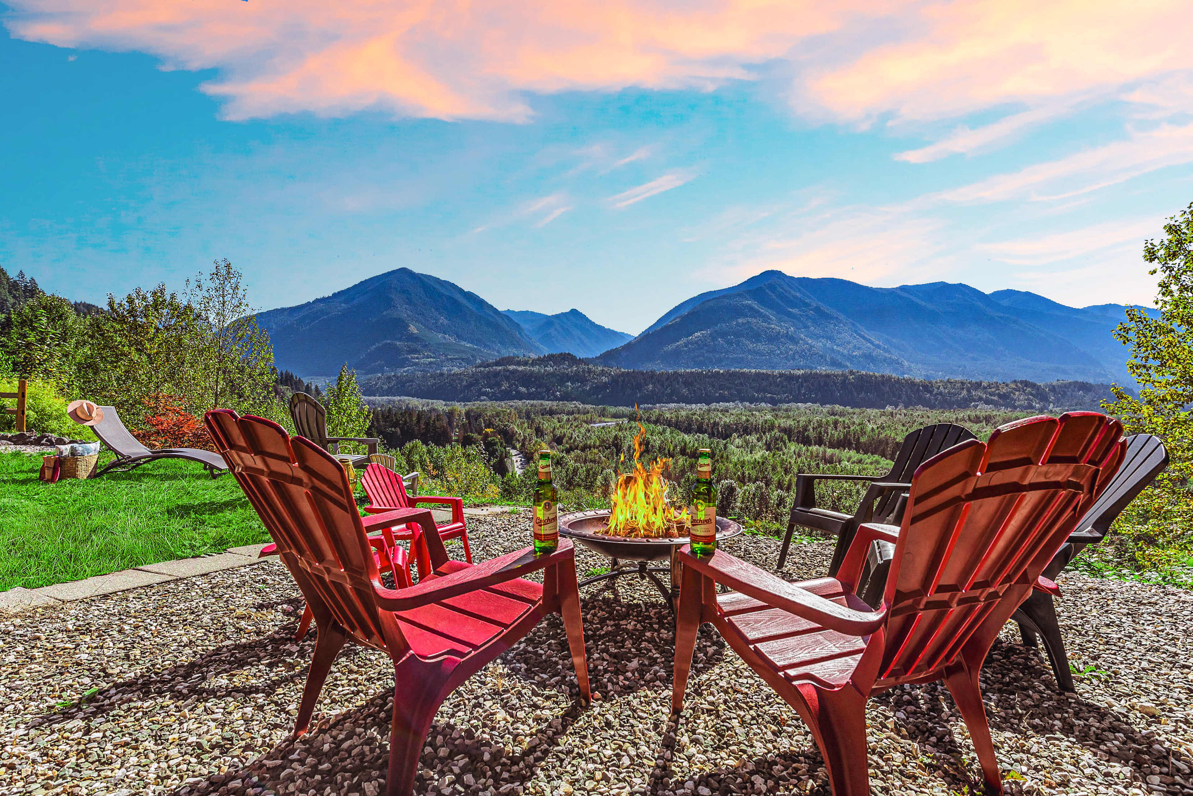 A circular arrangement of red Adirondack chairs surrounds a fire pit, with mountains and a colorful sky in the background.