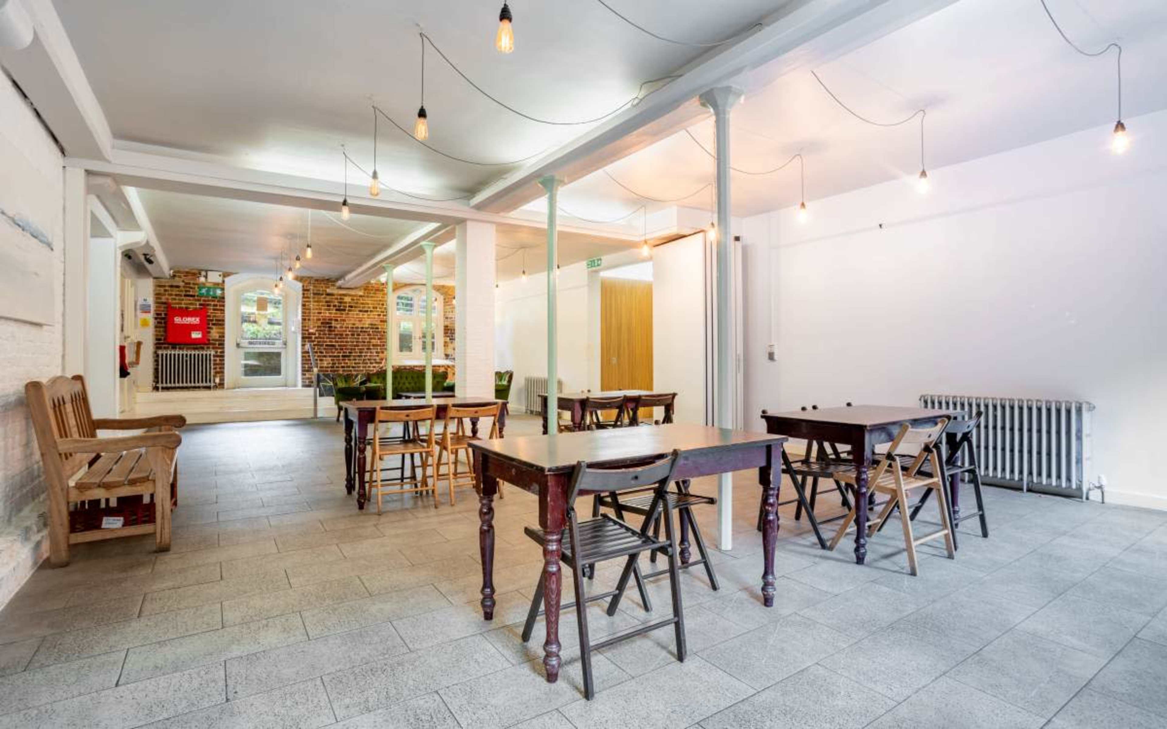 A spacious, well-lit interior with wooden tables and chairs arranged around a tiled floor, featuring hanging light bulbs and a few plants in the background.
