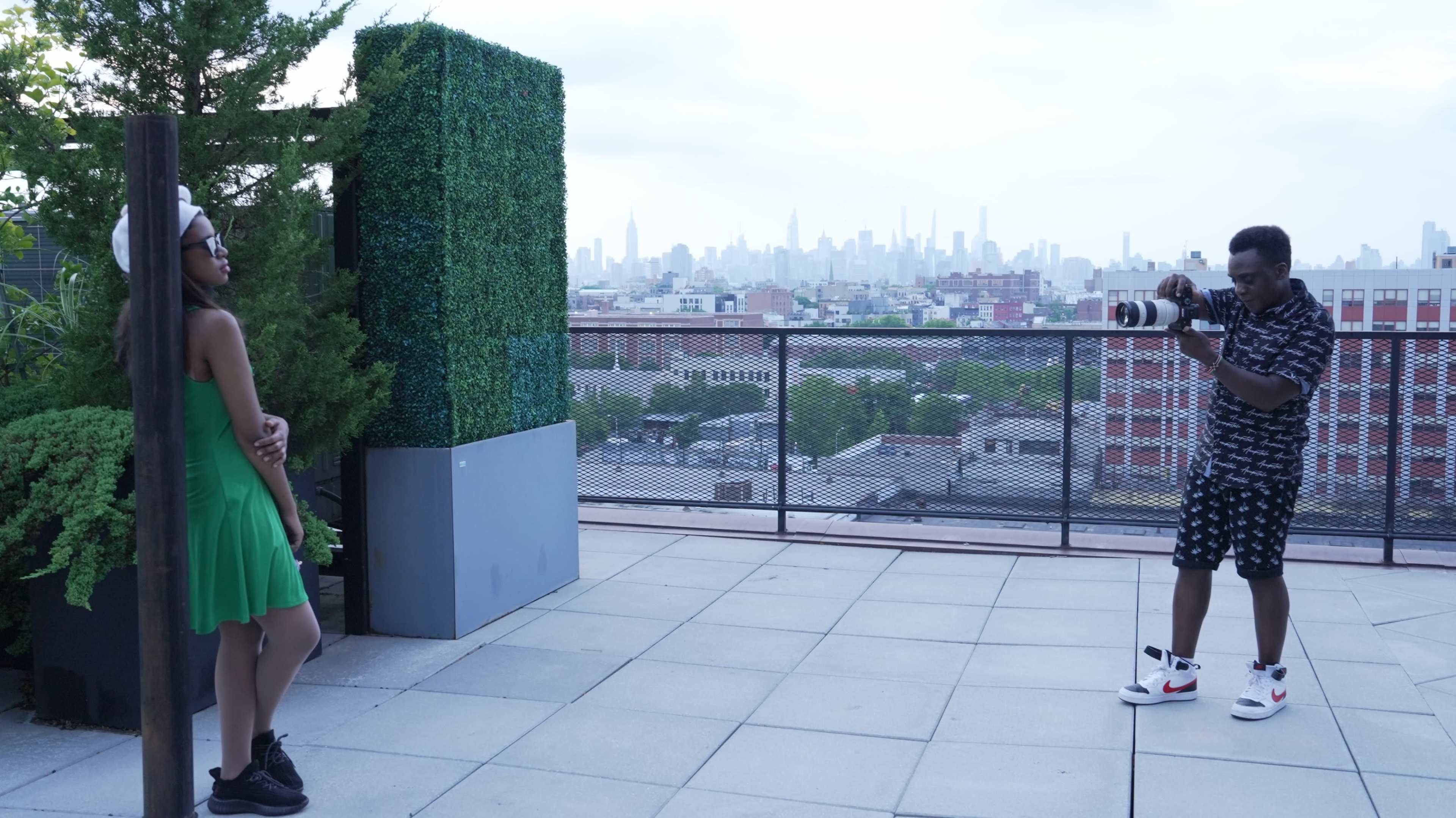 A photographer captures a model posing on a rooftop terrace with a city skyline in the background.