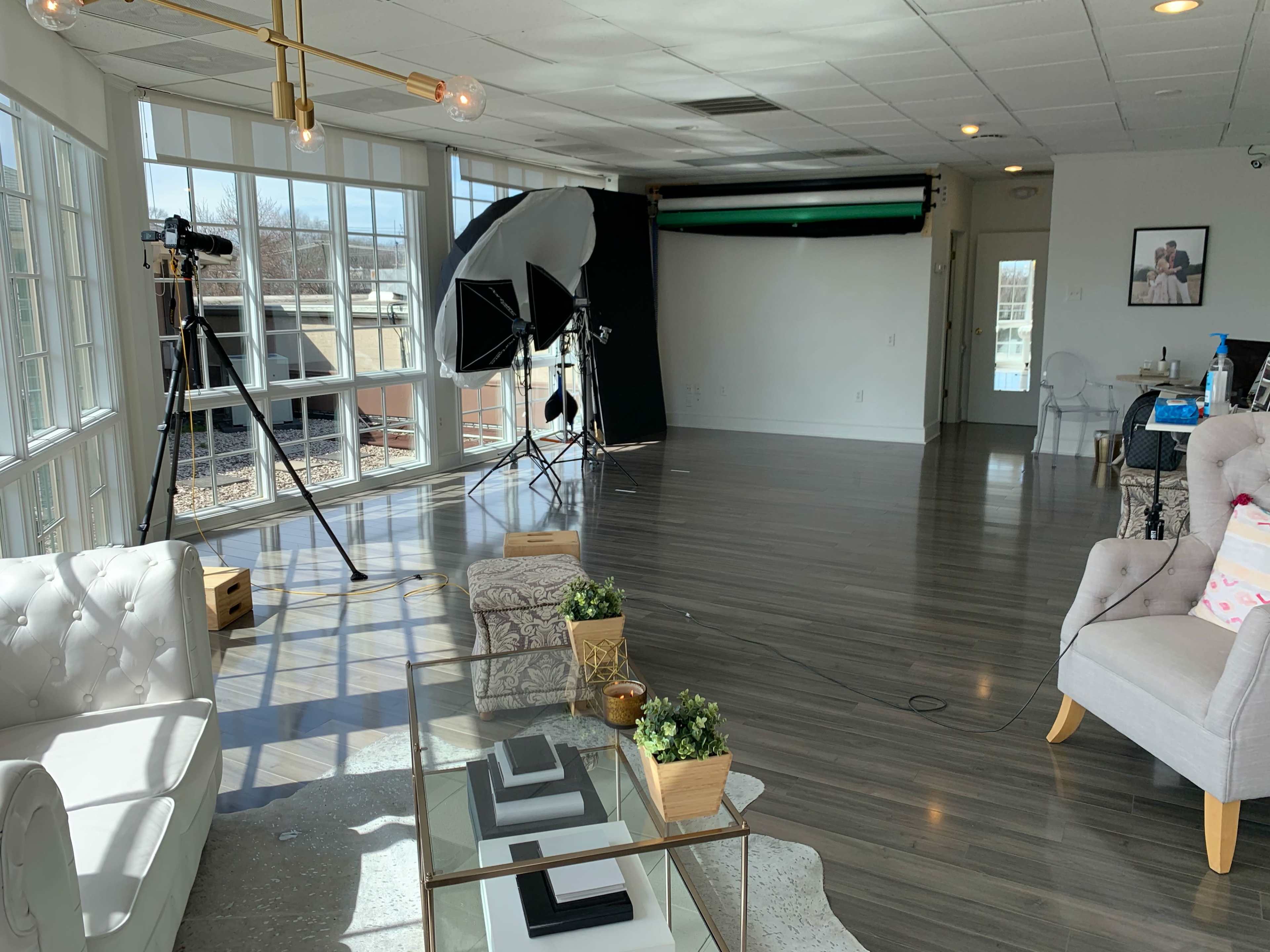 The image shows a well-lit studio space featuring a comfortable seating area, photography equipment, and a backdrop setup against a wall of windows.