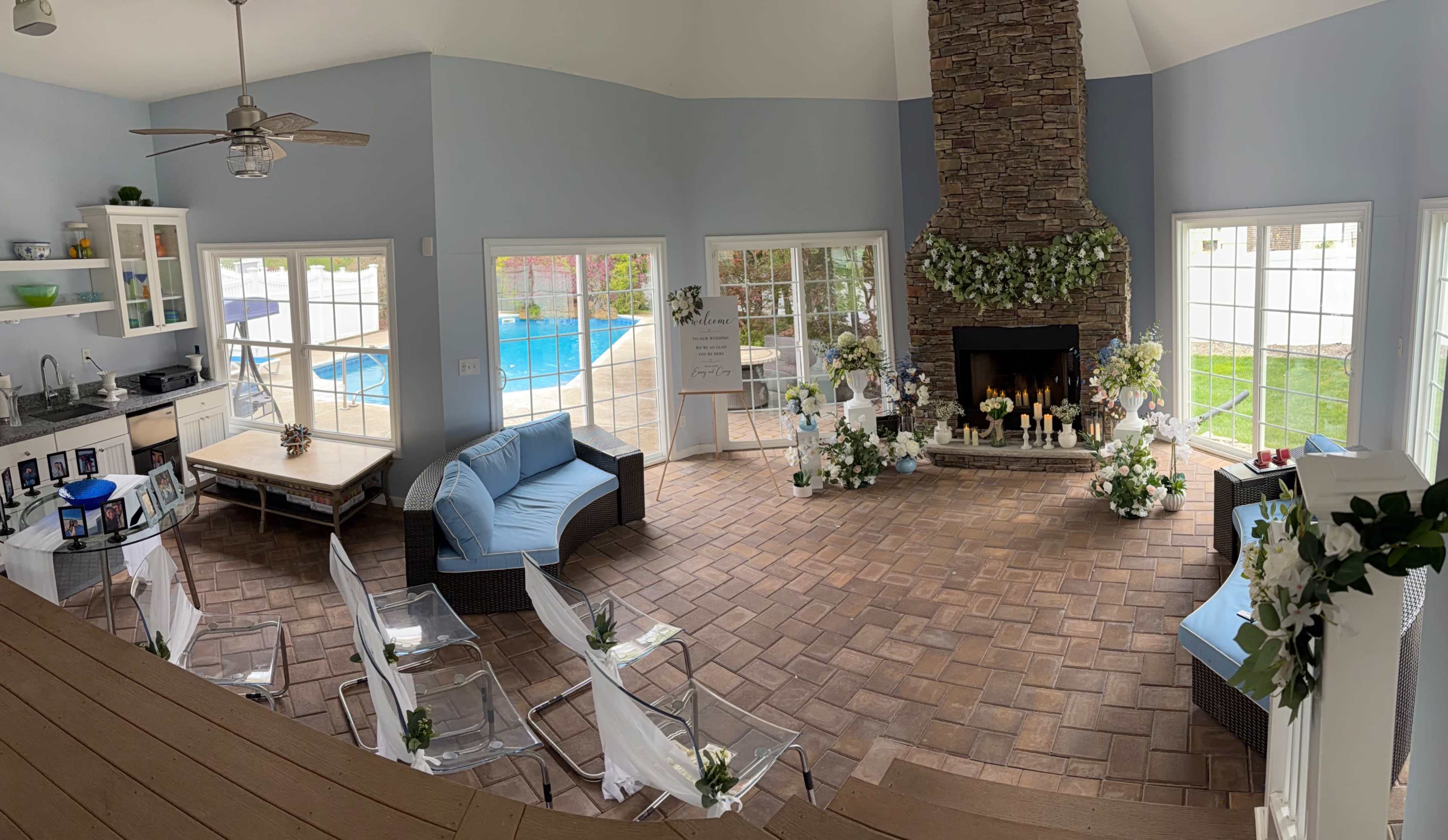 The image shows a spacious, light-filled room with a stone fireplace, seating arrangements, and large windows overlooking a pool area.