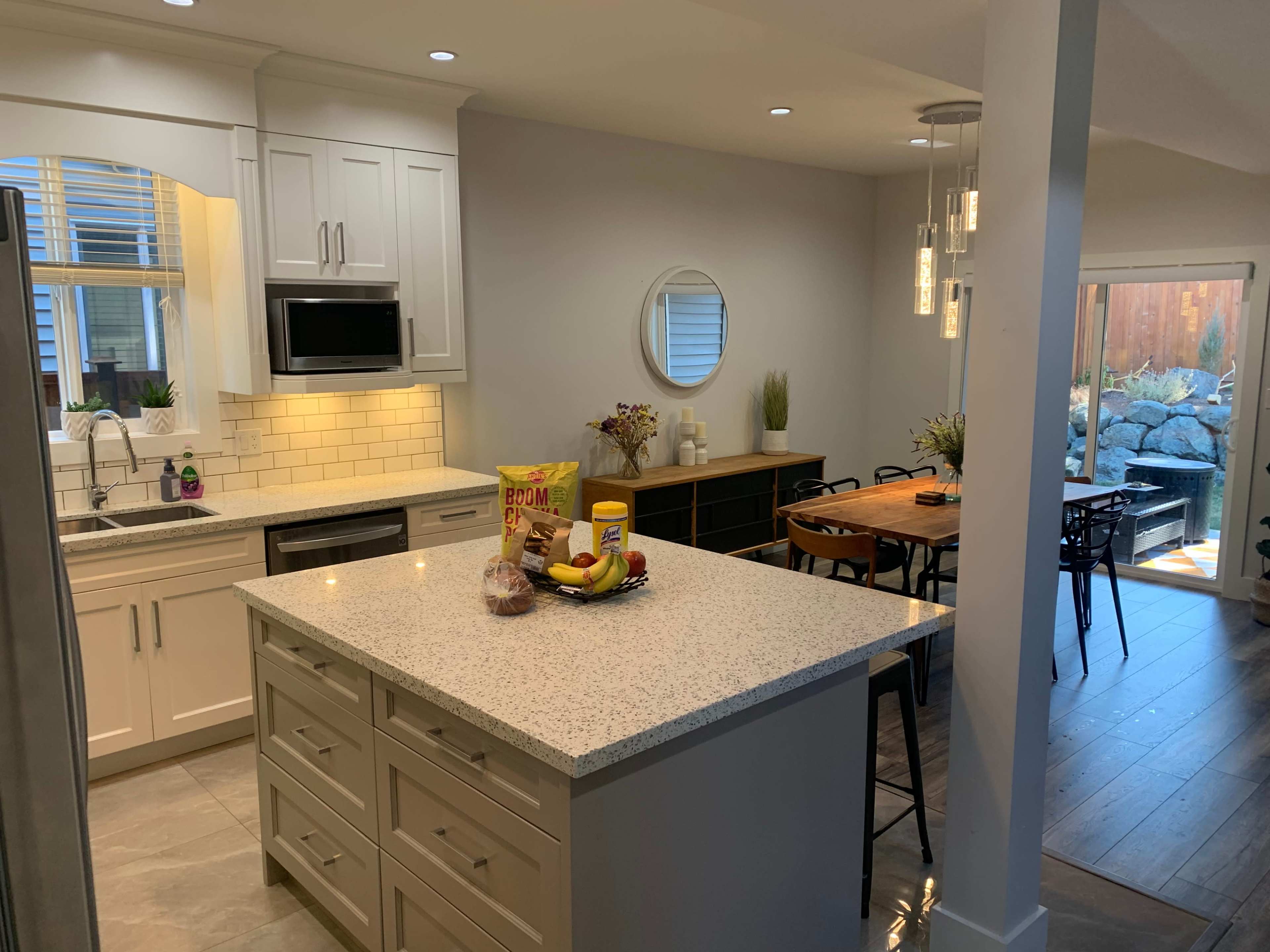A modern kitchen with a central island, featuring white cabinetry, a stainless steel refrigerator, and a dining area with wooden furniture and large windows.