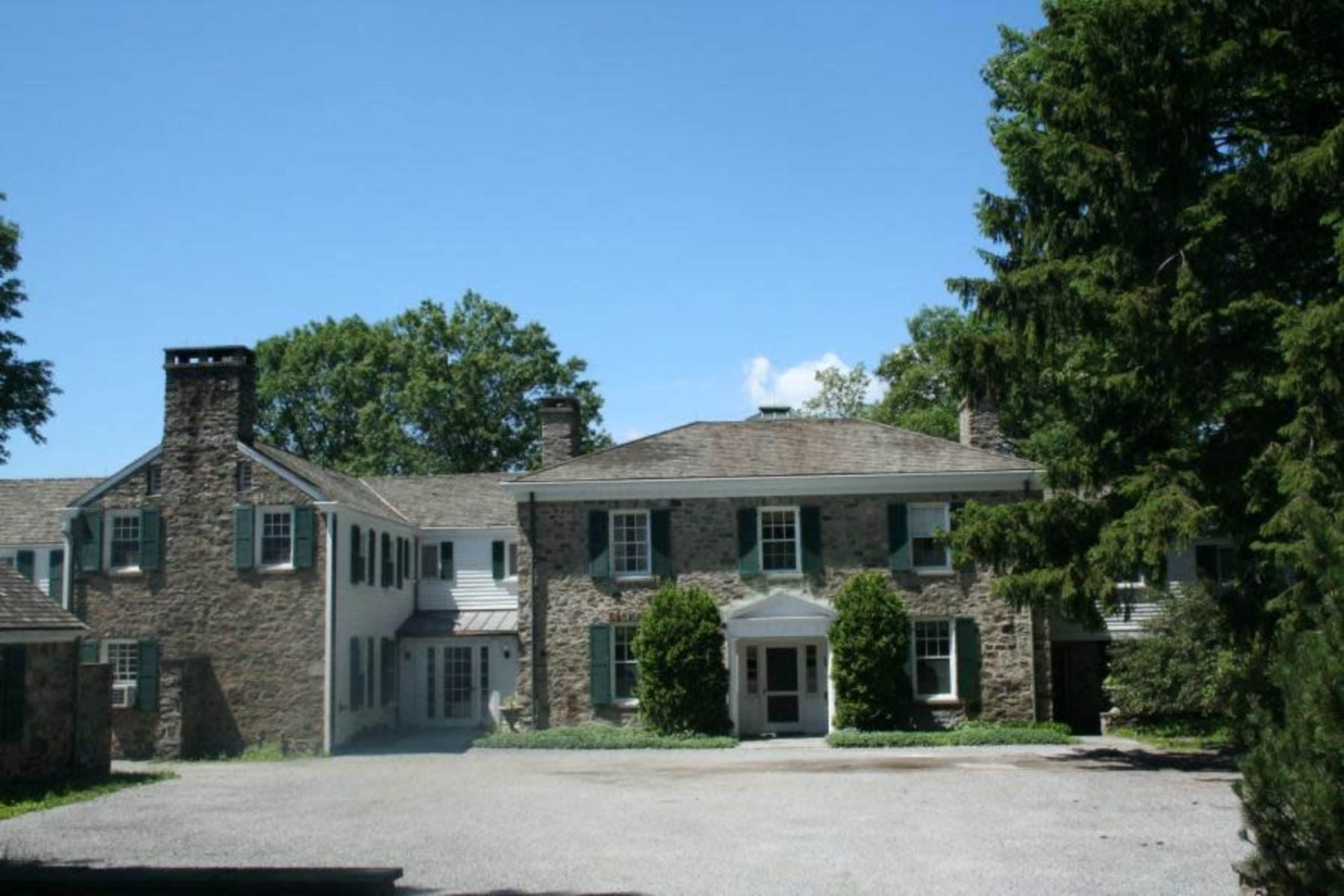The image shows a large stone house with multiple sections, surrounded by trees and a gravel driveway under a clear blue sky.