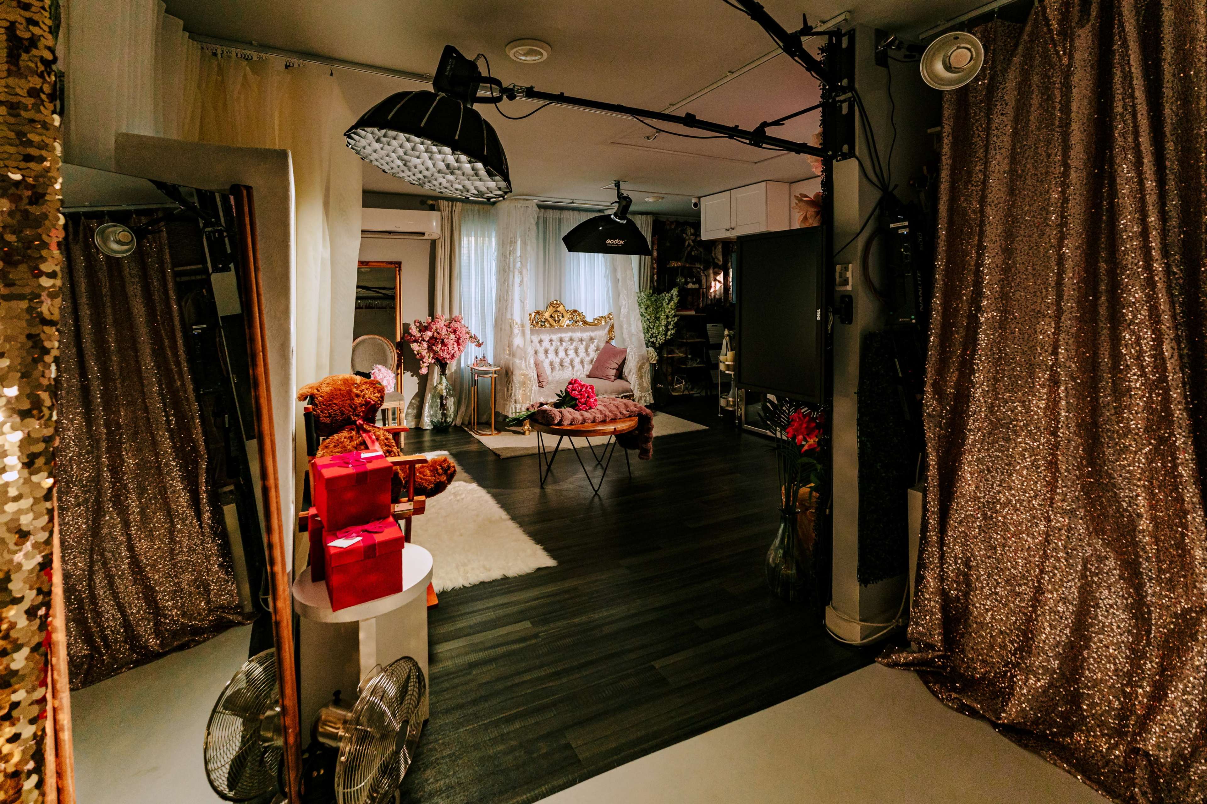 The image shows a stylish studio space featuring a combination of decorative curtains, lighting set-ups, and a cozy seating area with floral arrangements and gifts.