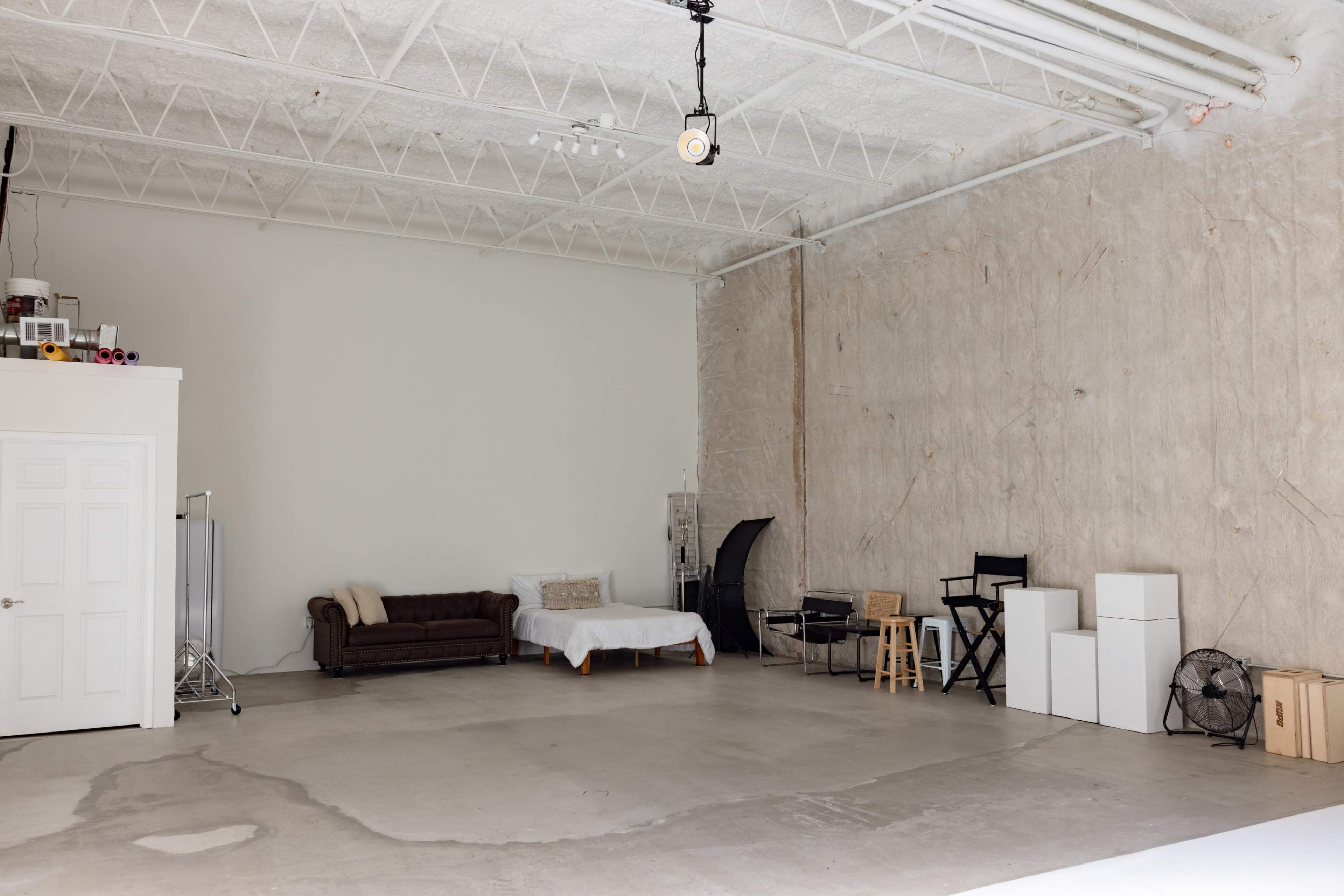 The image shows a spacious, empty studio with a couch, a bed frame without a mattress, and various other furniture items against a textured wall.