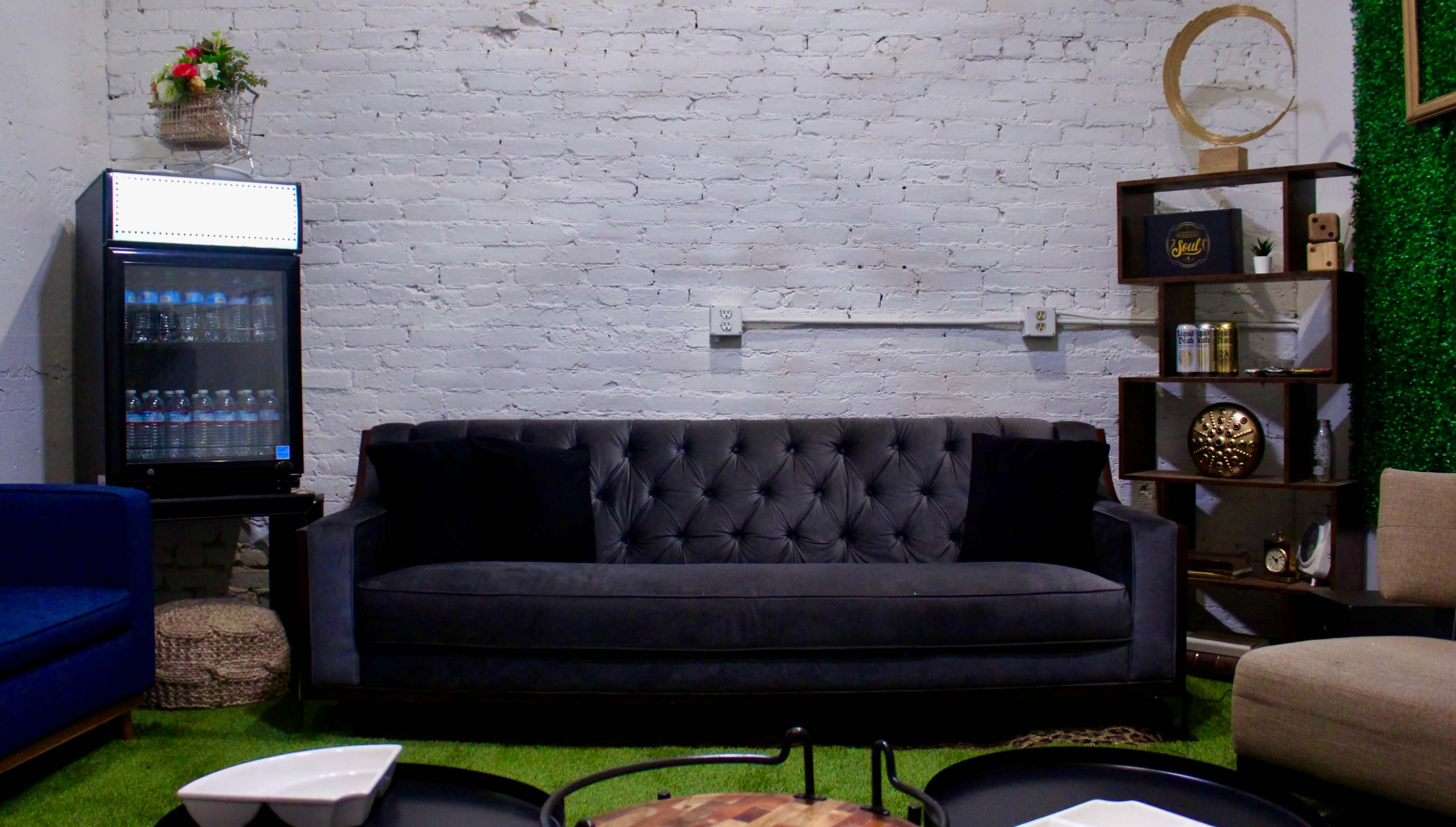 A living space featuring a dark tufted sofa against a white brick wall, a glass-fronted refrigerator, wooden shelves, and green grass-like carpet.