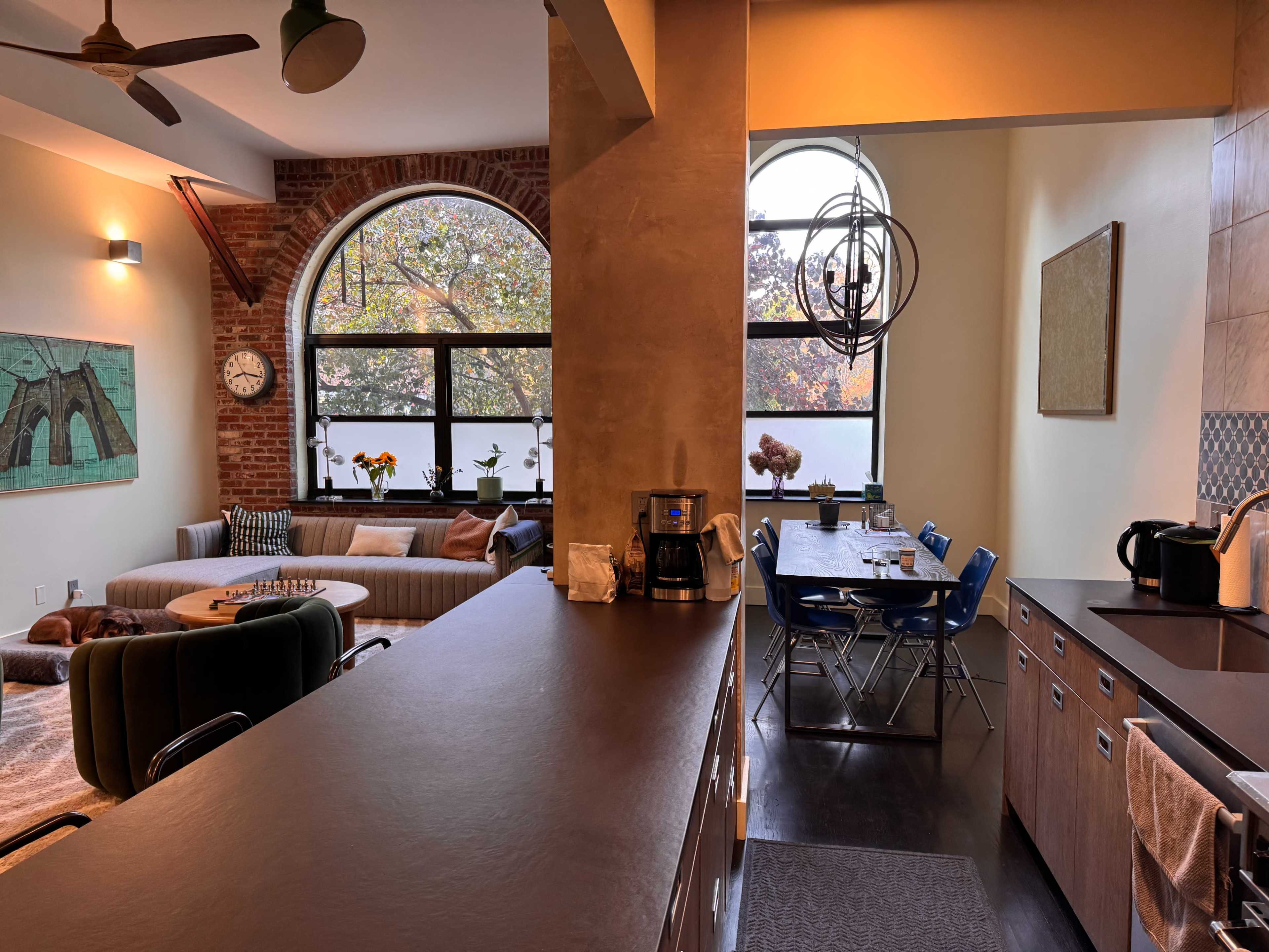 The image shows a modern kitchen and living space with large windows, exposed brick walls, a dining table with blue chairs, and a comfortable seating area.