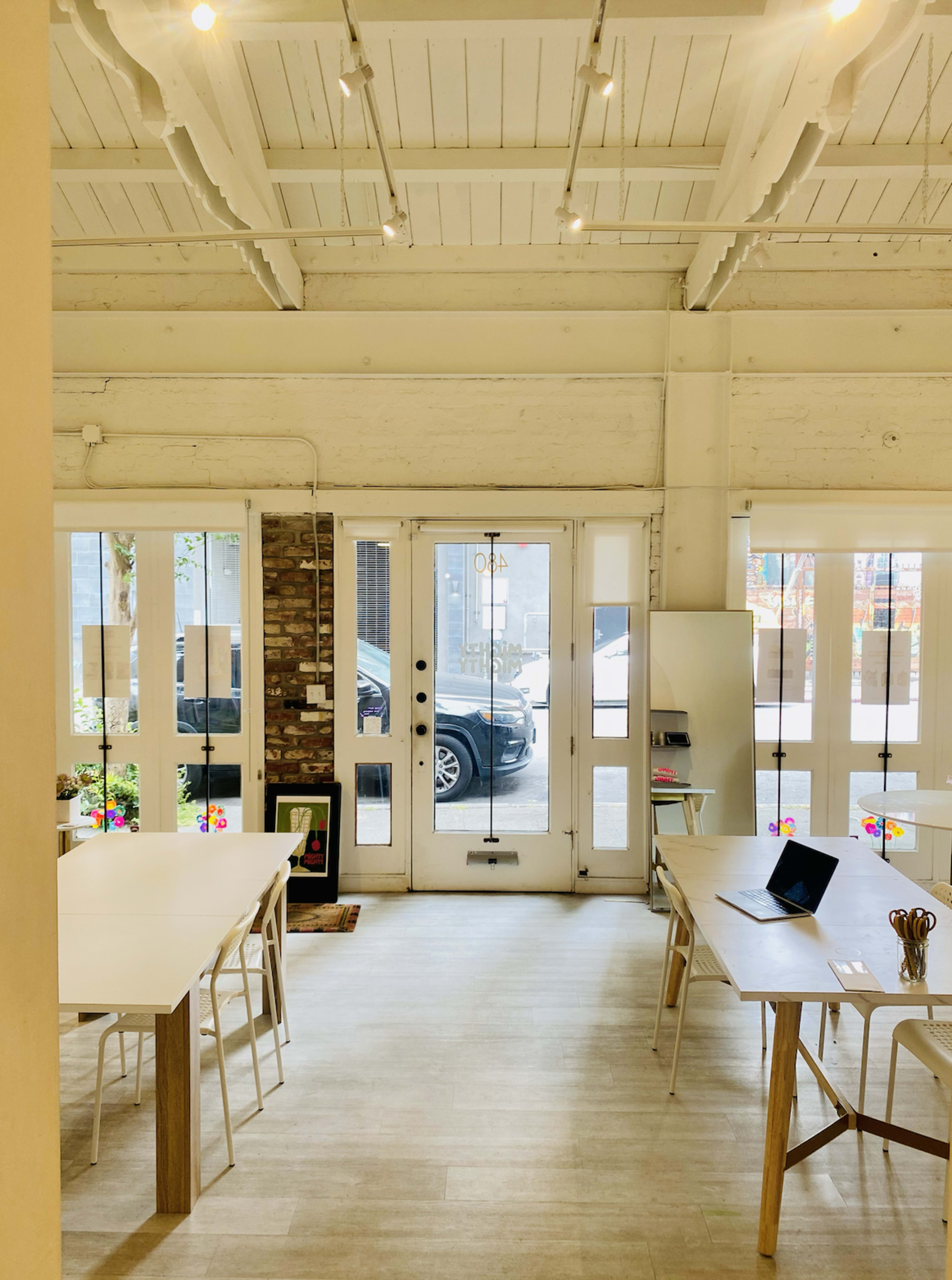 The scene features a bright, open workspace with white furniture, wood beams, and large glass doors leading to an outdoors area.