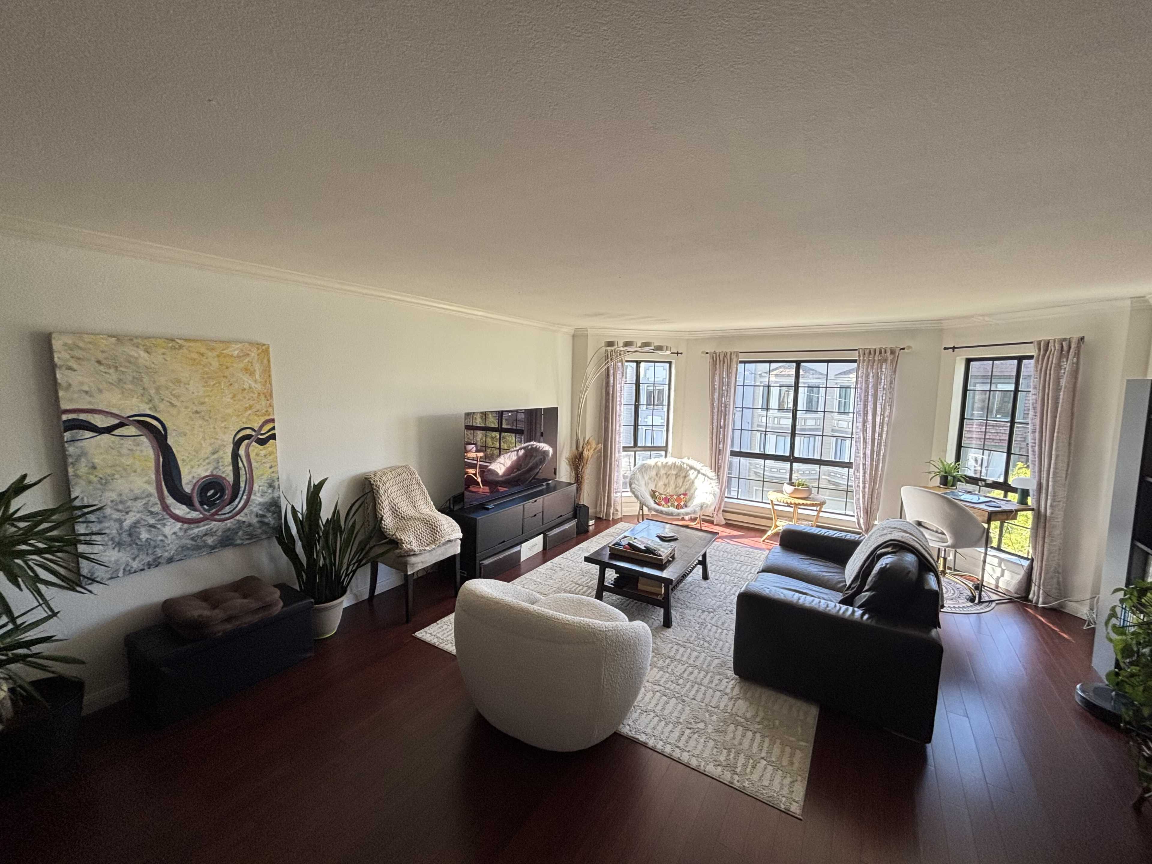 The image shows a spacious living room with wooden flooring, a large television, a sofa, an armchair, and windowed walls allowing natural light to enter.