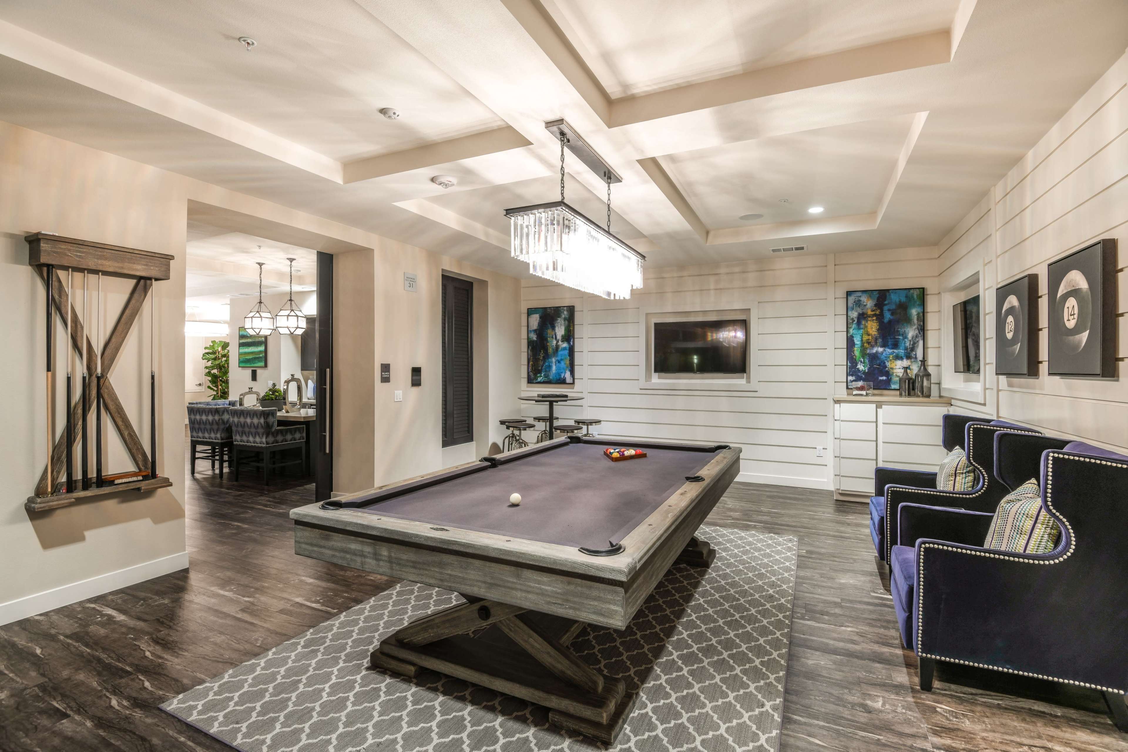 The image shows a game room featuring a billiards table, two blue armchairs, and a dining area visible through an open doorway.