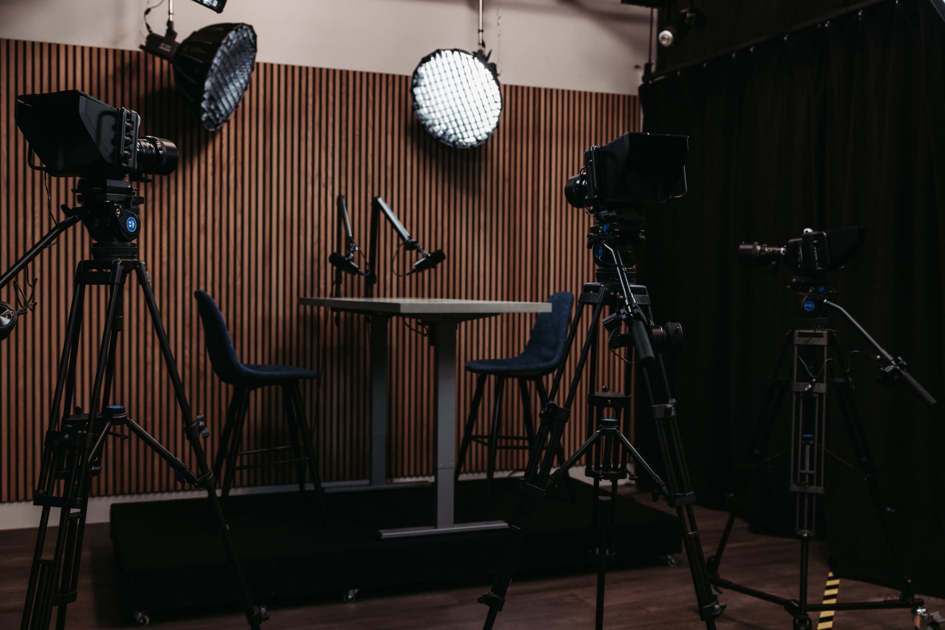 A studio setup features two high chairs and a table, surrounded by several cameras and bright lights on a wooden-paneled backdrop.