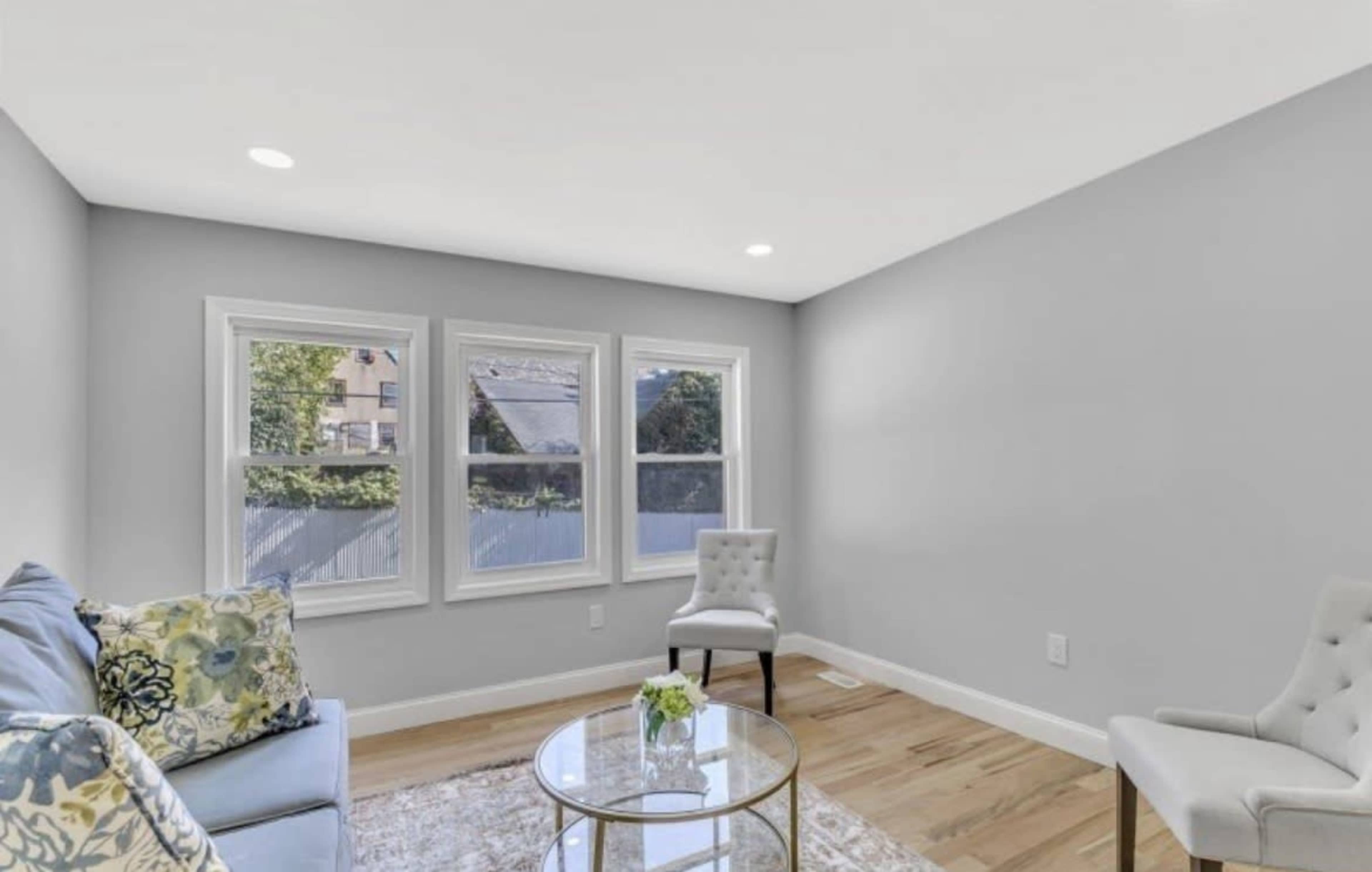 The image shows a light gray living room featuring a blue couch with decorative pillows, two chairs, a round glass table, and three windows revealing a view outside.