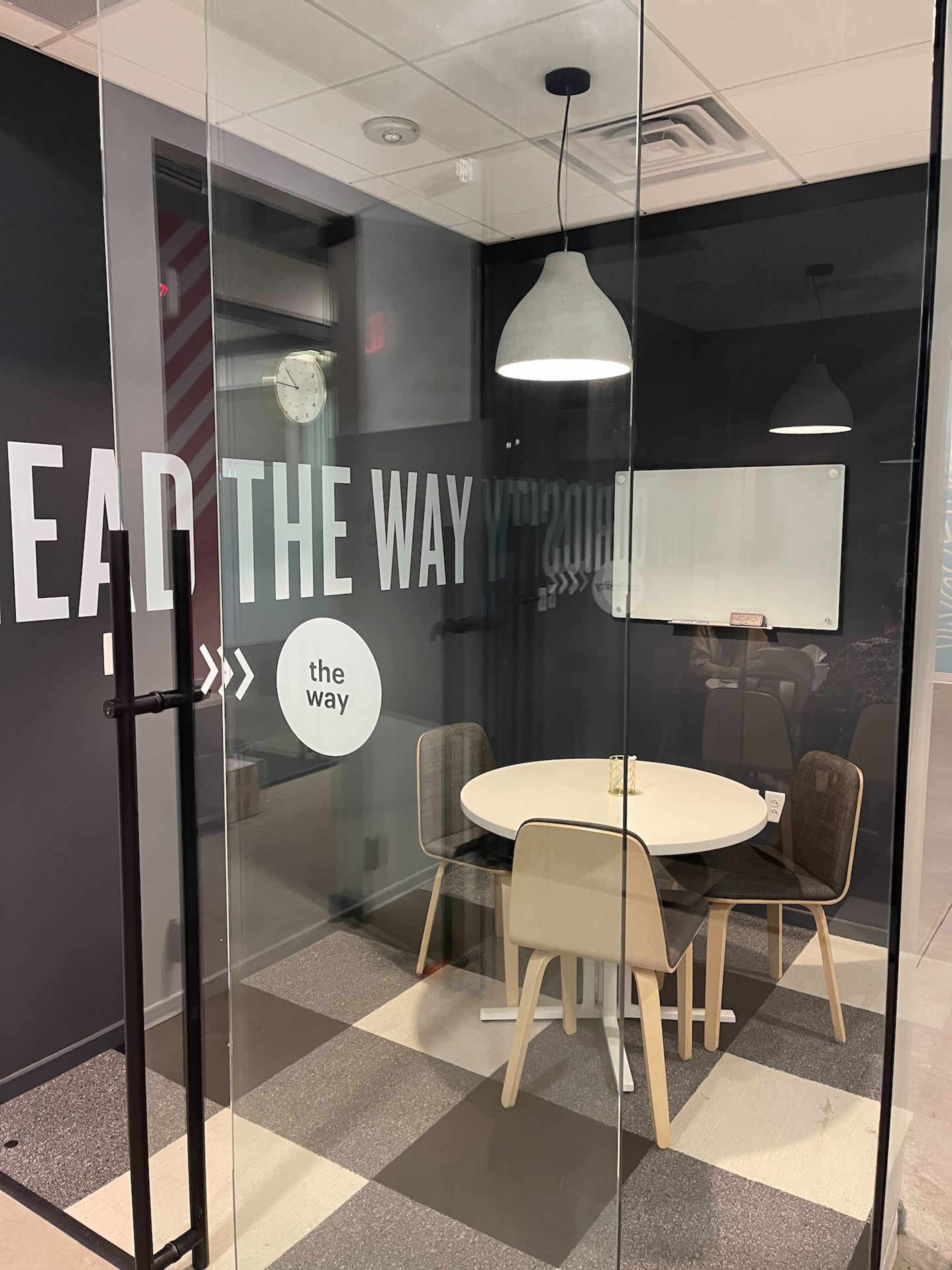 A glass-walled meeting room features a round table surrounded by four chairs, with a whiteboard and wall graphics.
