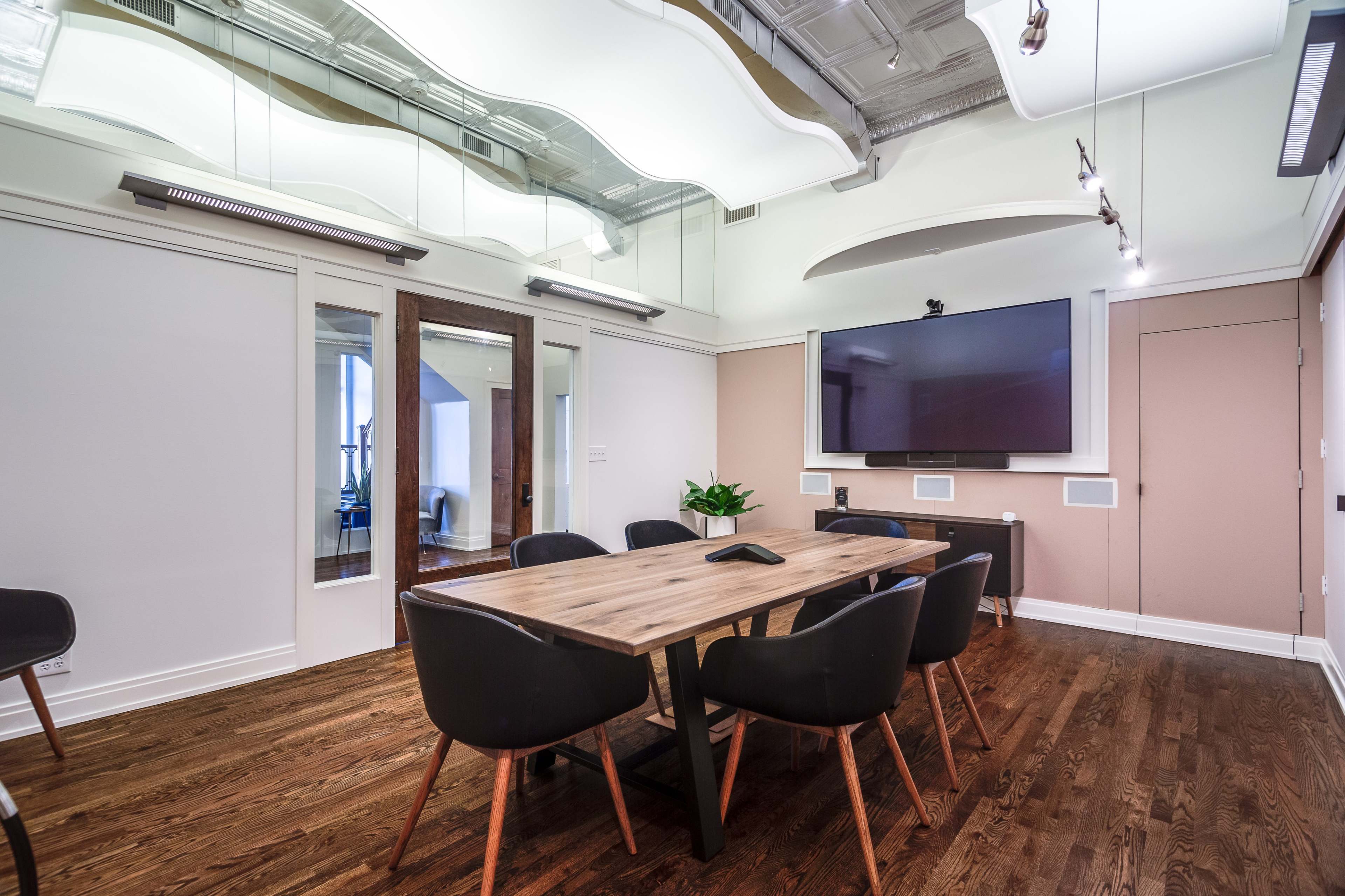 A modern conference room features a wooden table surrounded by black chairs, with a wall-mounted television and large windows allowing natural light.
