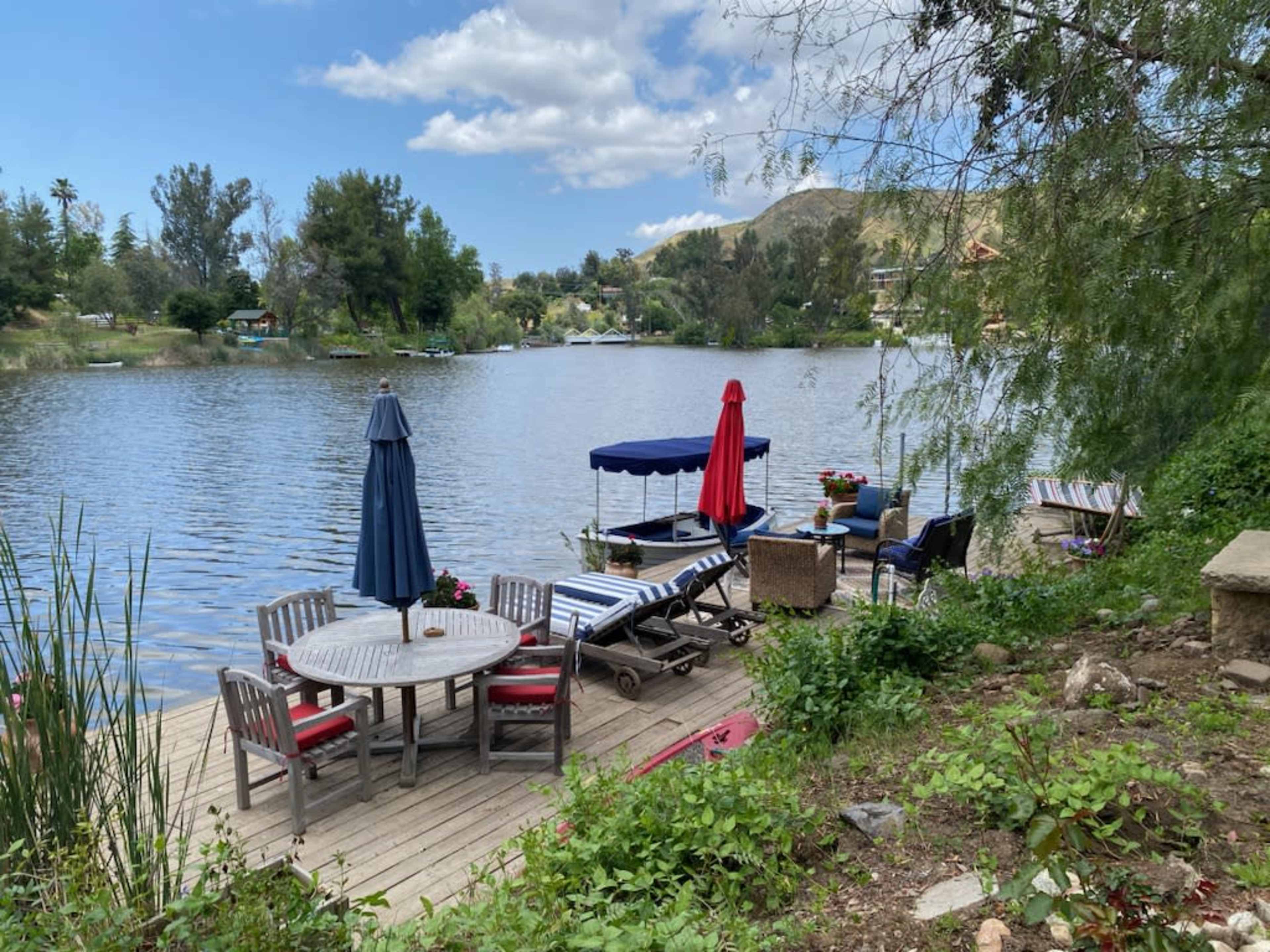 A wooden deck by a calm lake features a round dining table, lounge chairs, and umbrellas, surrounded by greenery and distant hills.