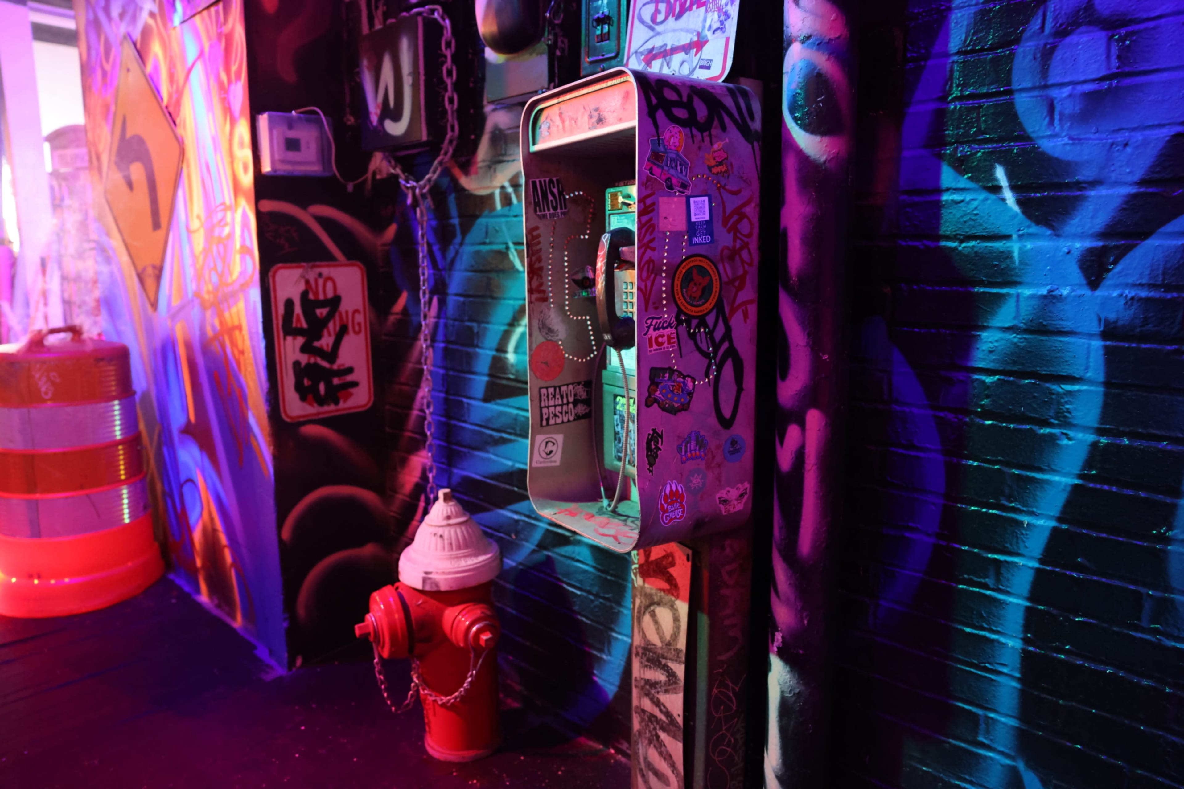 The image shows a decorated payphone beside a red fire hydrant, set against a backdrop of vibrant graffiti on a wall.