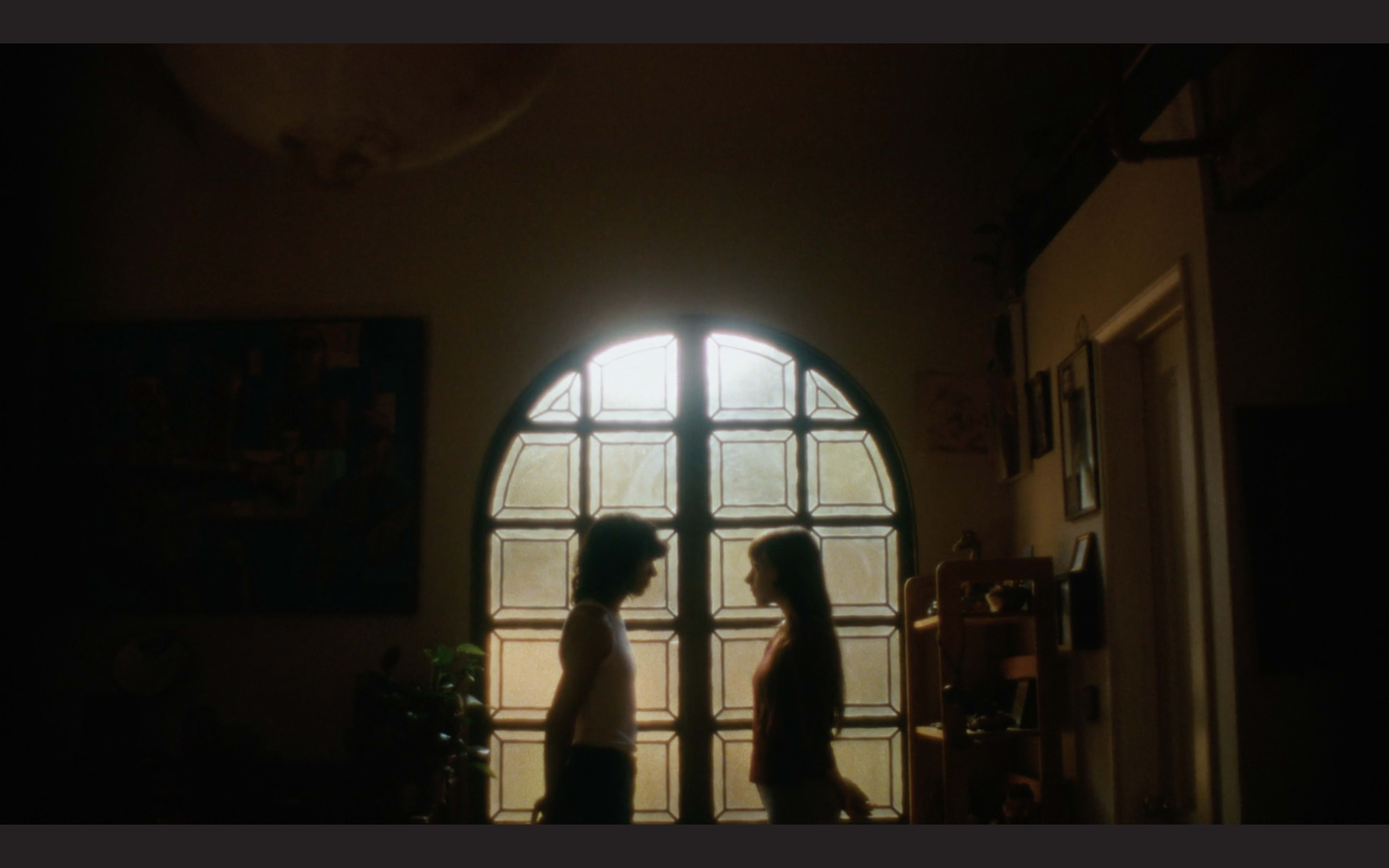 Two figures stand facing each other in front of a large window with a distinctive pattern of glass panes.