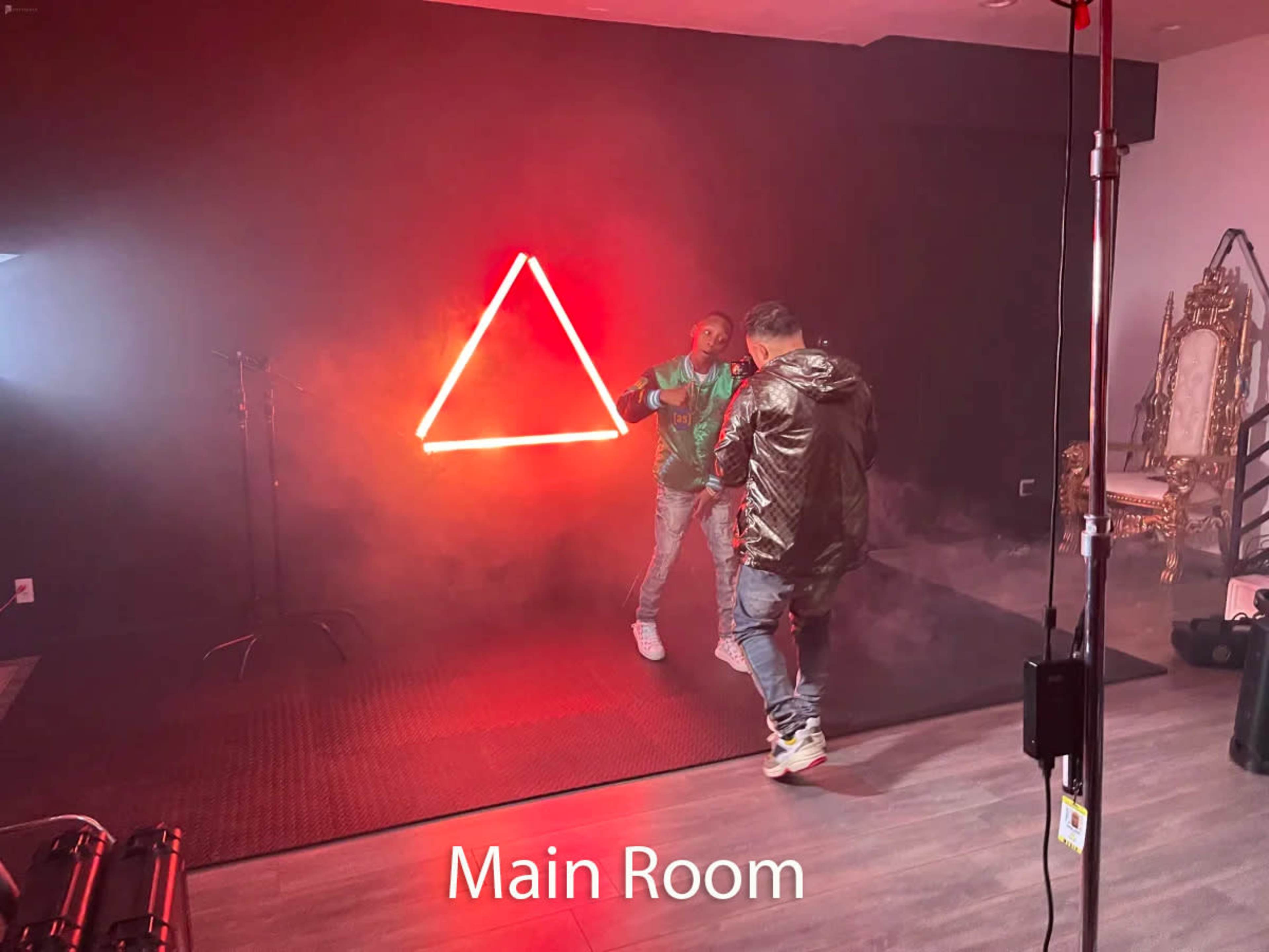 Two individuals are positioned in a dimly lit main room featuring a triangular neon light, with smoke effects creating an atmospheric backdrop.