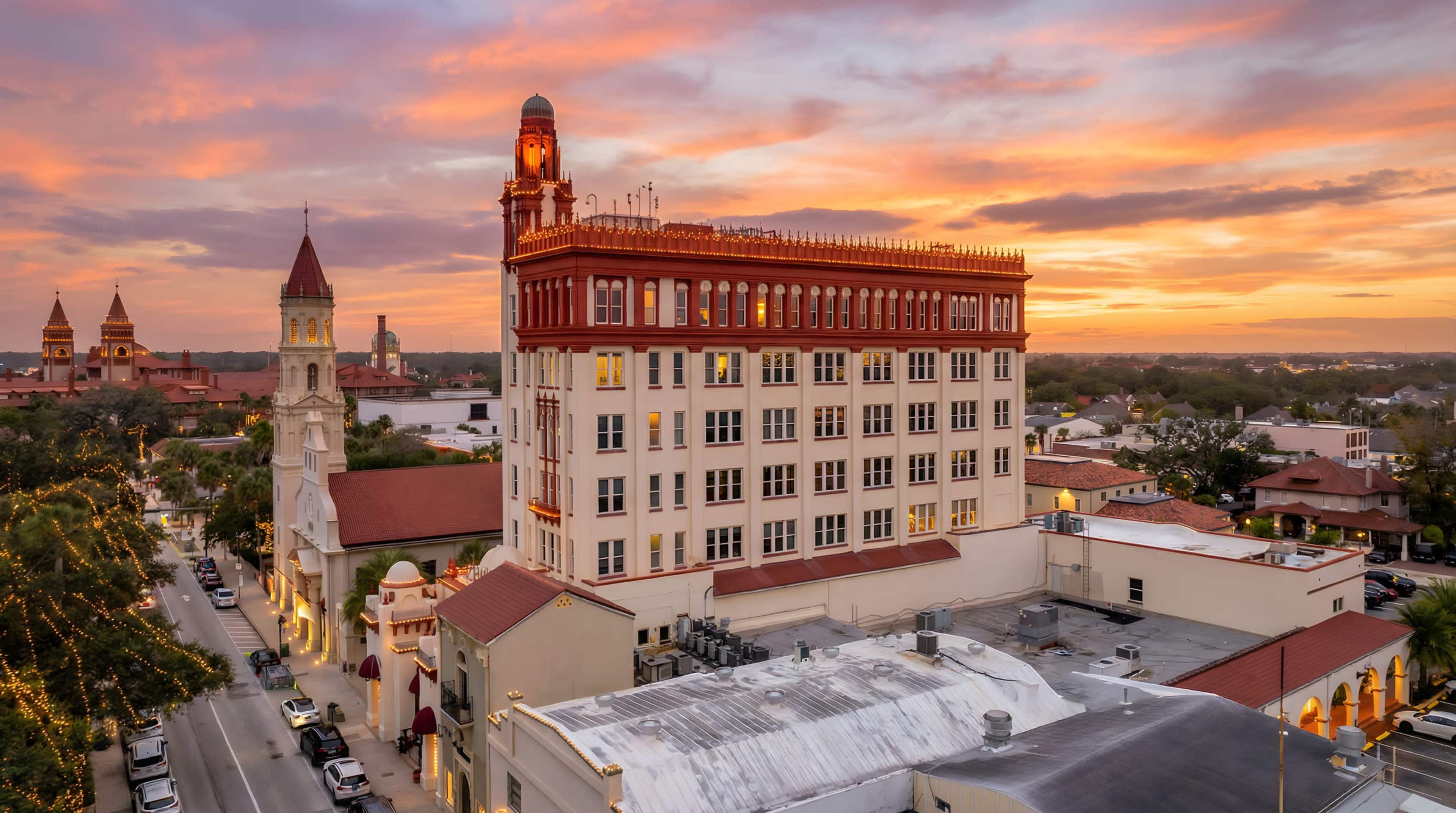 The image shows a historic building with a red and white facade against a vibrant sunset sky, surrounded by other structures and trees.