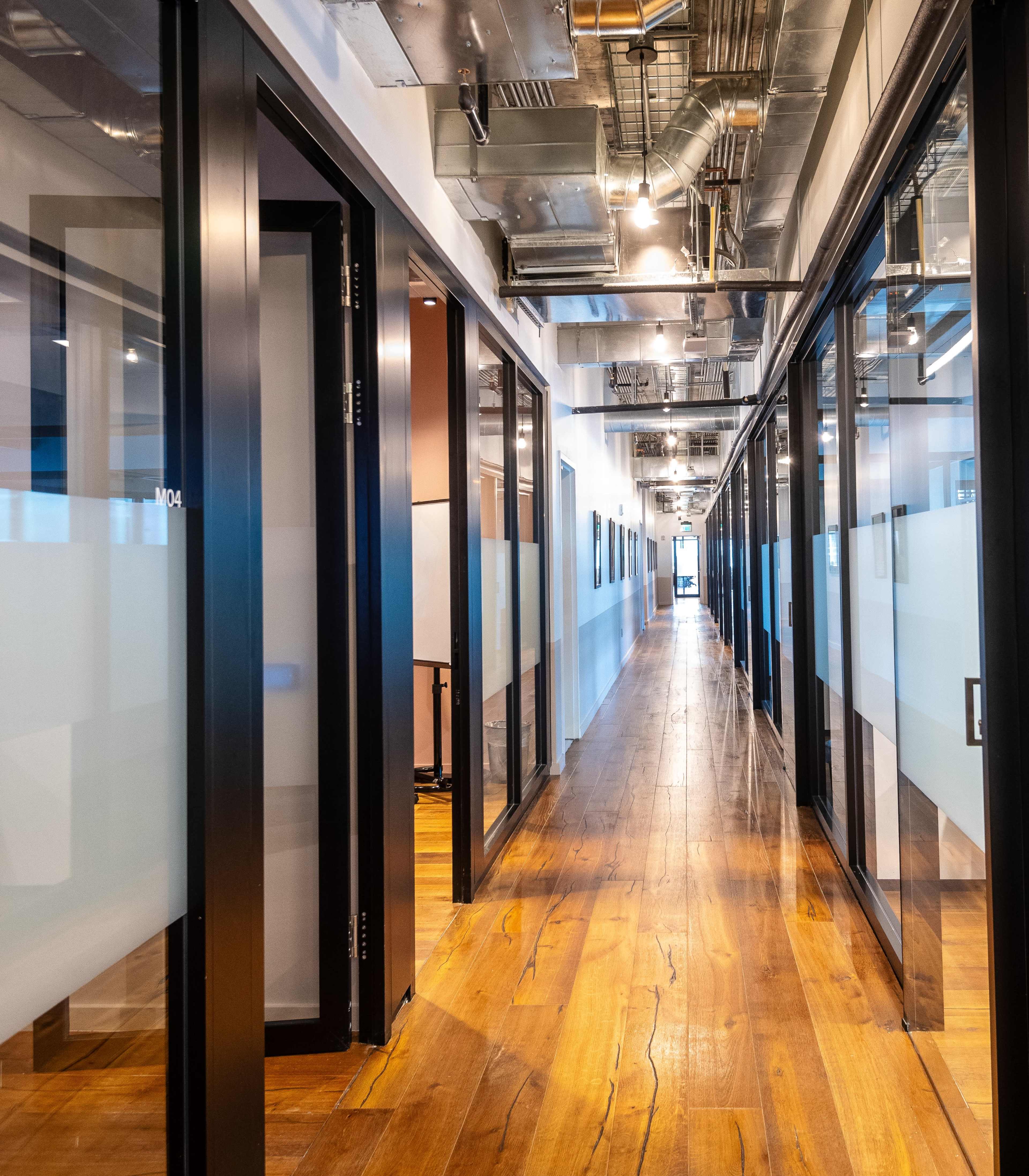 The image shows a long corridor lined with glass-walled office rooms and wooden flooring, illuminated by overhead lights.