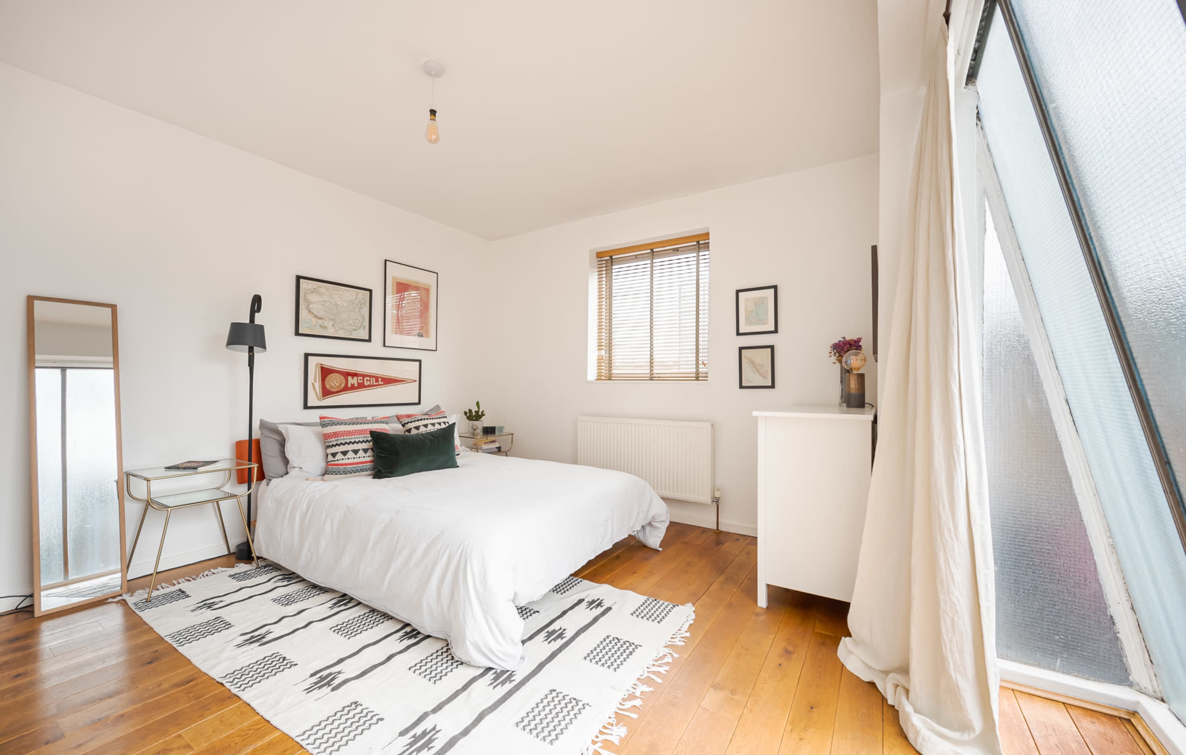 A bright bedroom features a white bed, wooden flooring, framed artwork on the walls, and a large window with blinds.