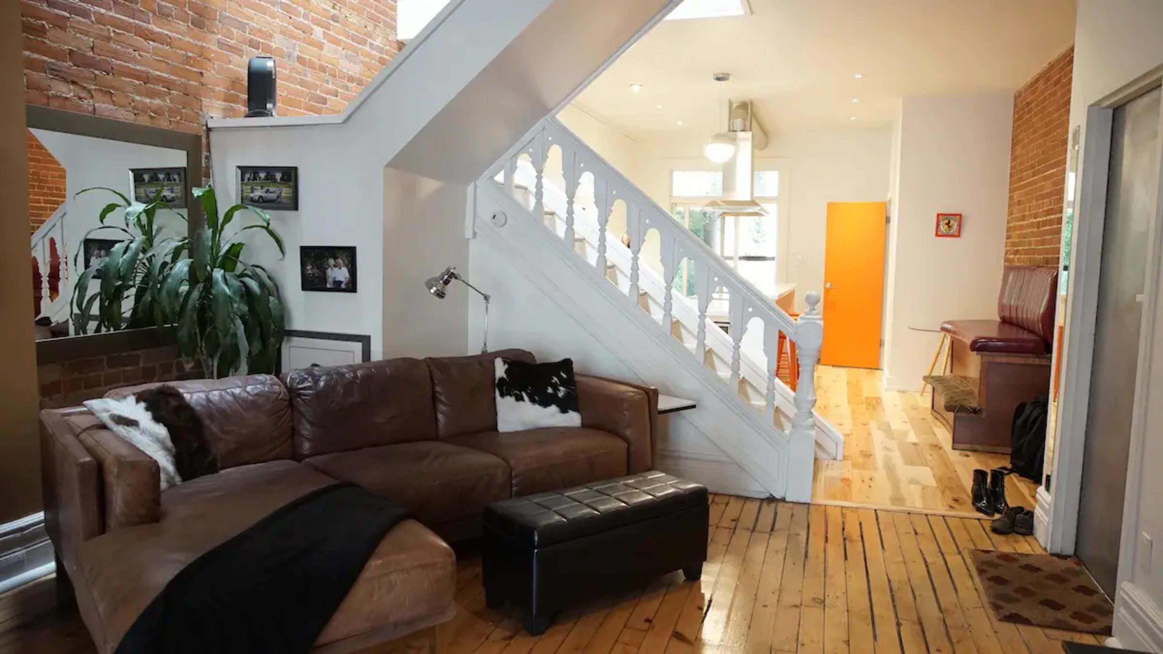 The image shows a well-lit living room with a brown leather couch, a black ottoman, and a staircase leading to an upper level, with a view of a hallway and an orange door.