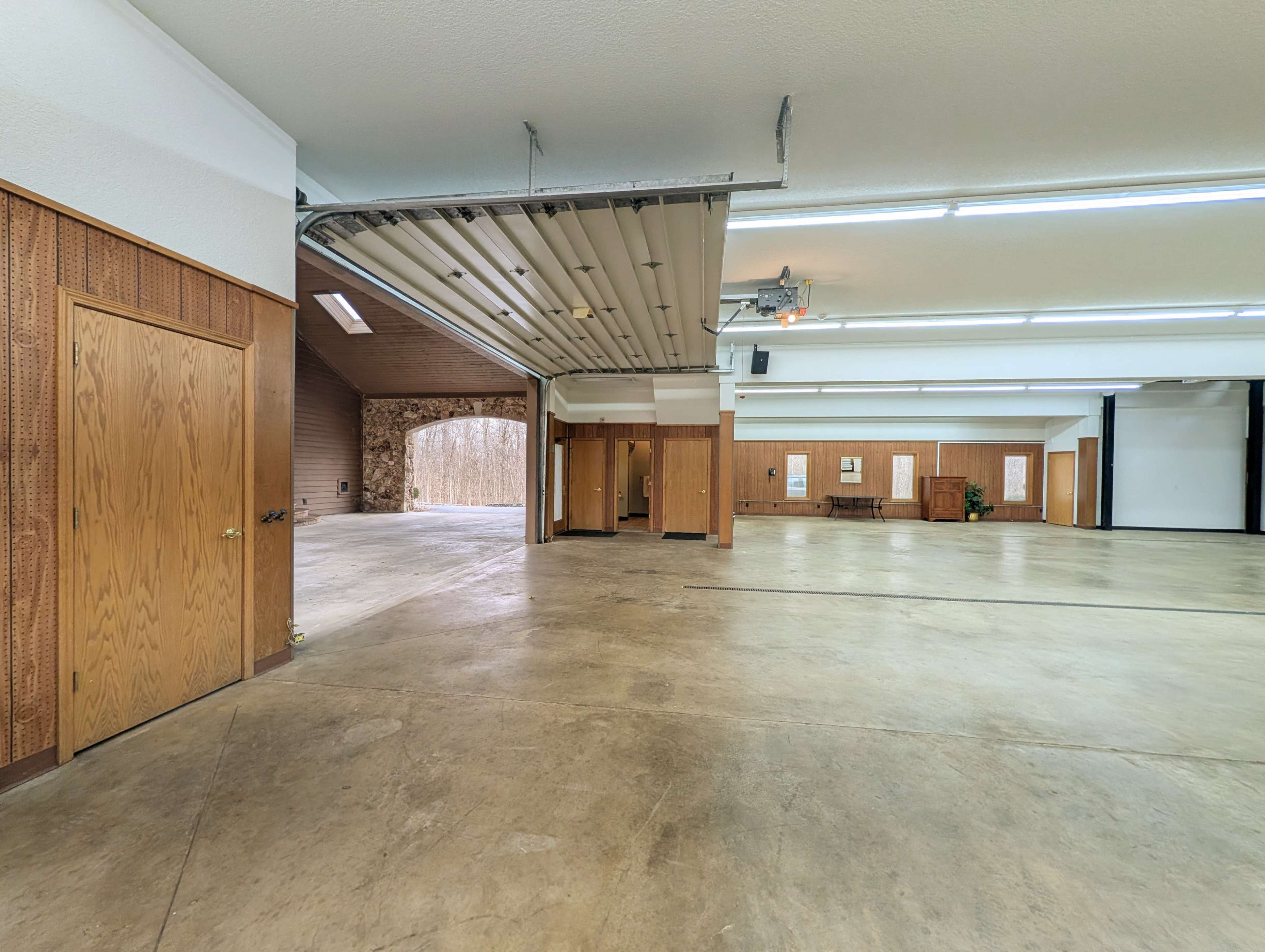 The image shows a spacious, empty garage with concrete flooring and a high ceiling, featuring a large garage door and wooden accents on the walls.