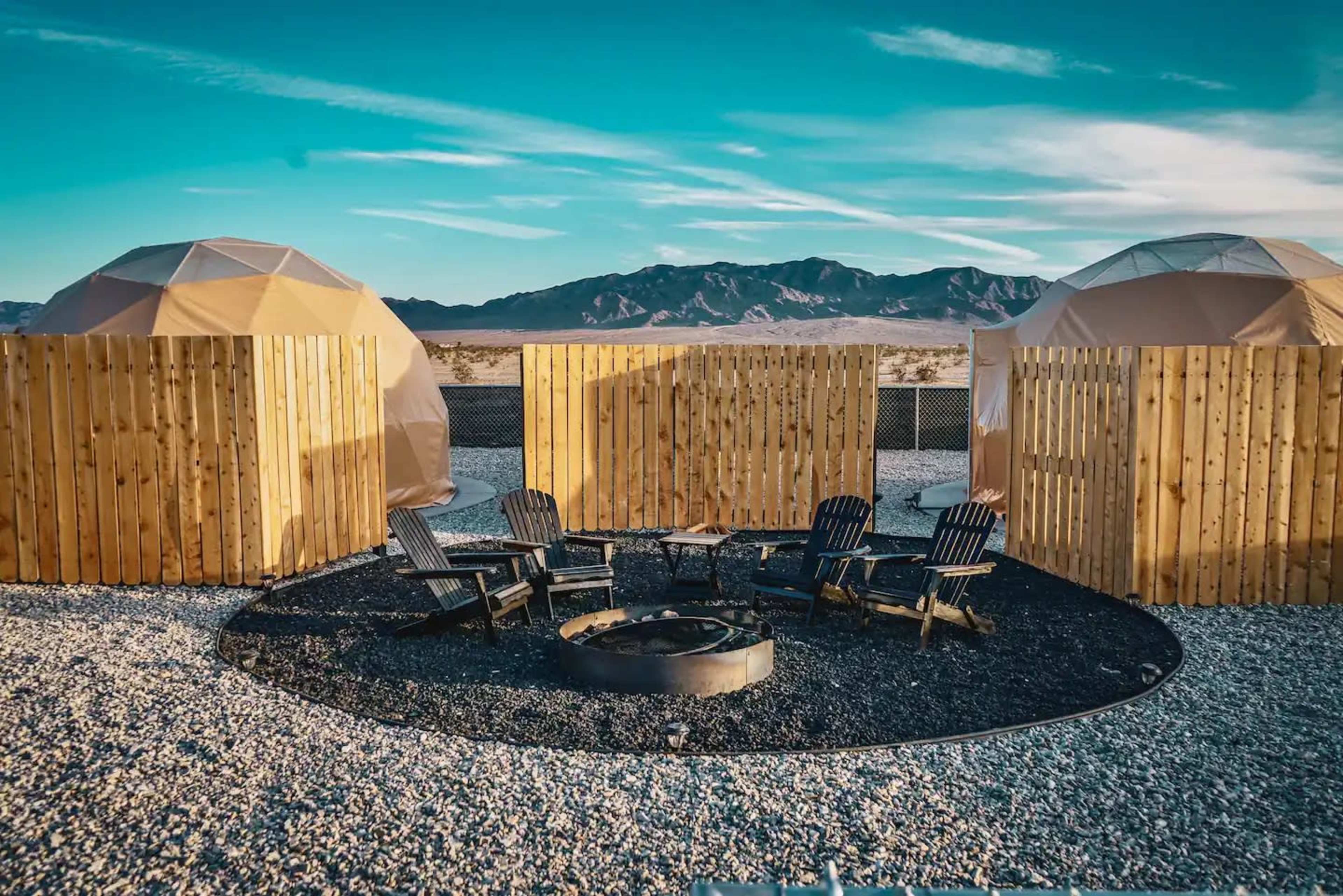 The image shows a group of wooden chairs surrounding a fire pit in a gravel area, with geodesic domes and wooden fences in the background under a blue sky.