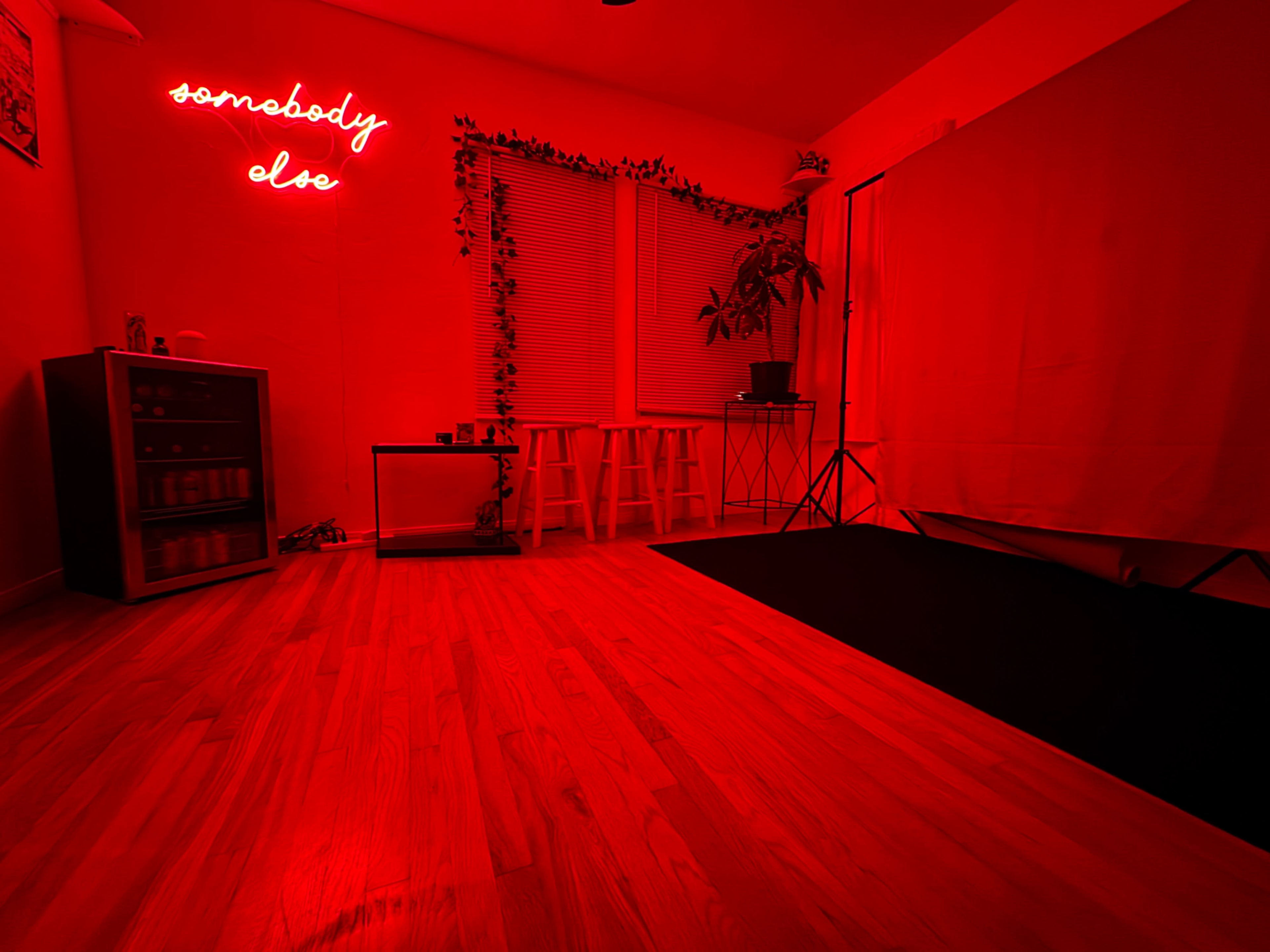 The room features a red neon sign saying "somebody else," wooden flooring, and minimal furnishings including a cabinet and two stools.