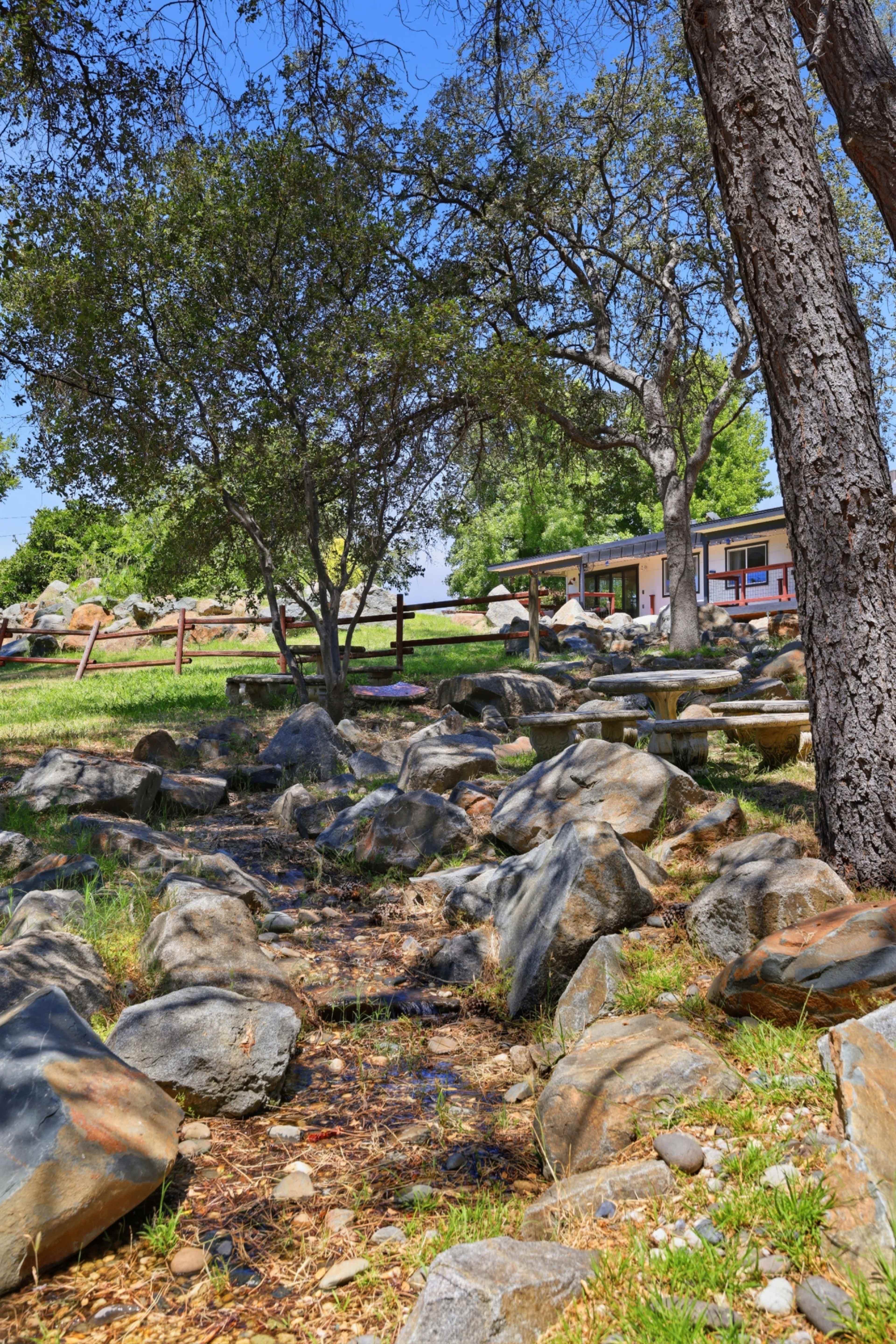 A rocky stream flows through a grassy area with trees and a building in the background.