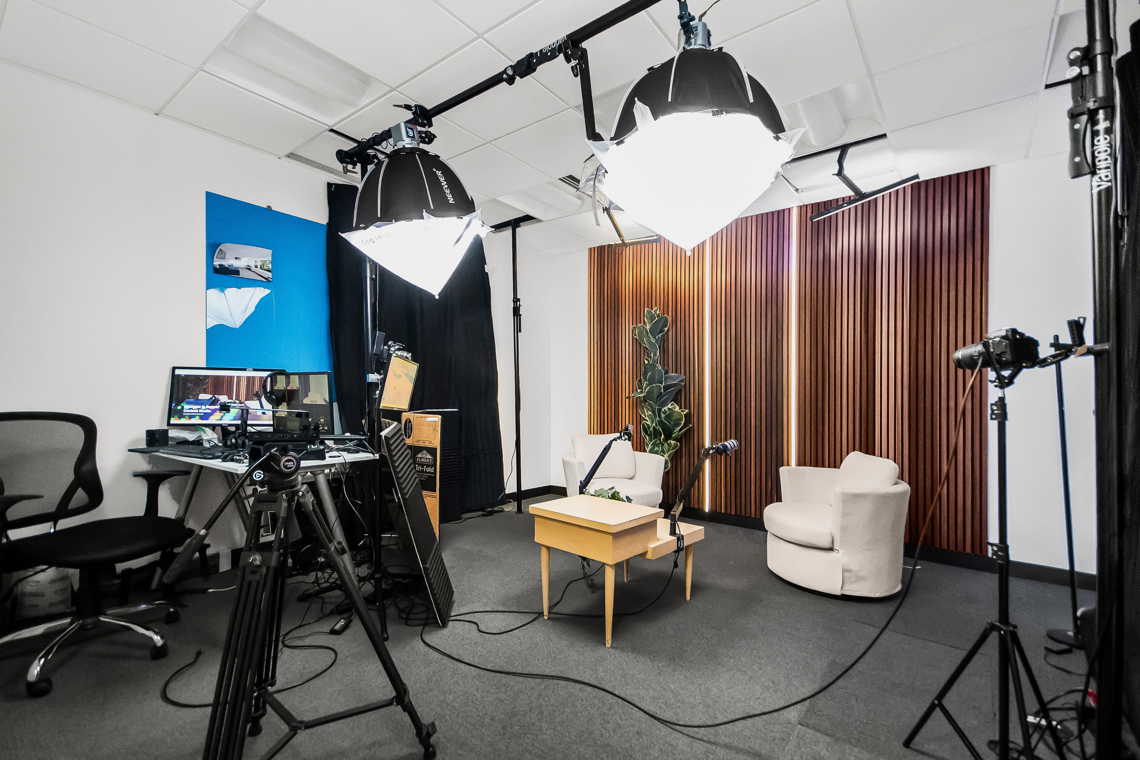 A video recording studio is set up with two large lights, a camera on a tripod, a microphone, a small table with a plant, and a white chair against a backdrop of wood paneling.