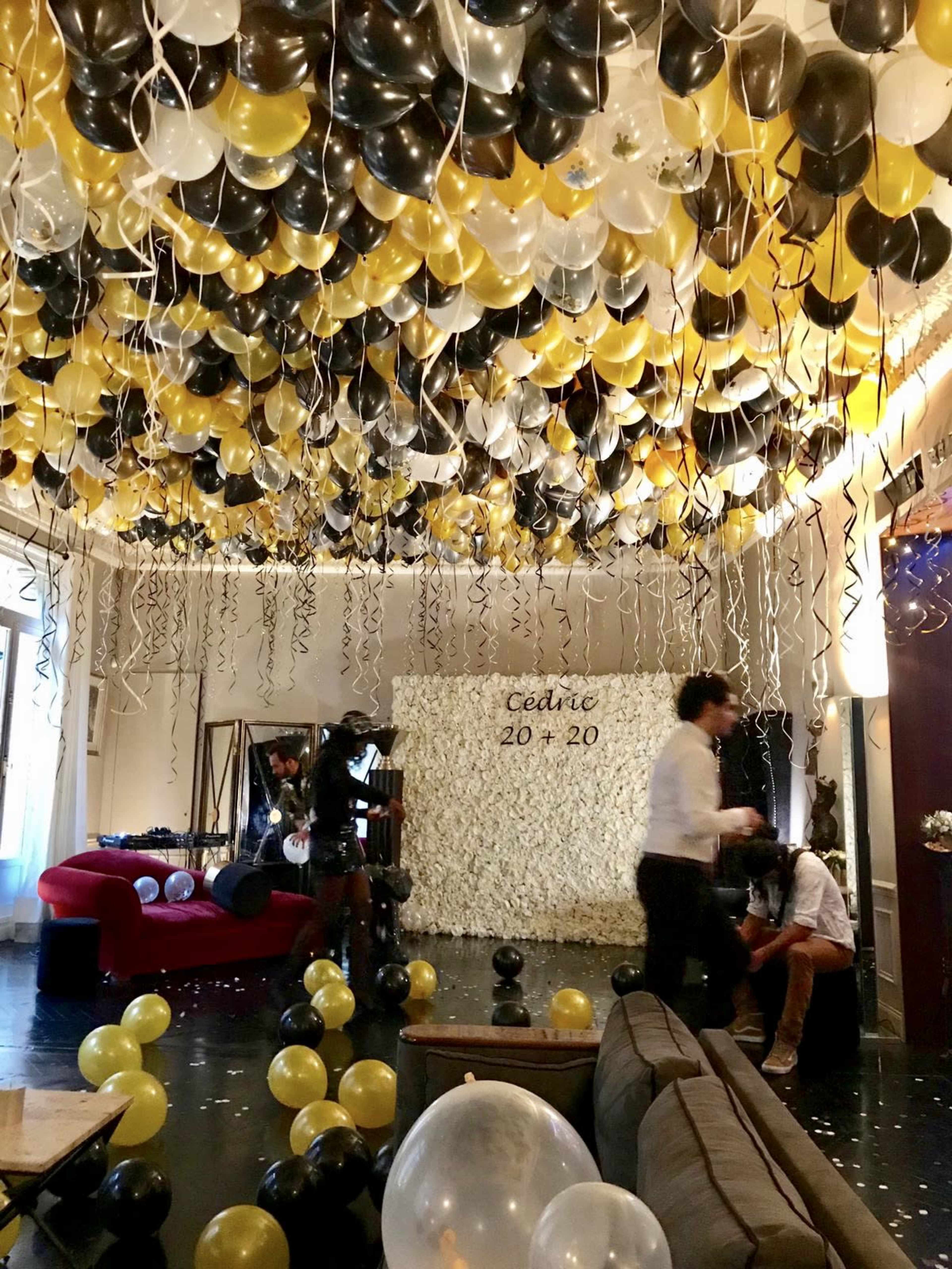 A party room is decorated with a ceiling full of black, gold, and white balloons, with a floral backdrop featuring the message "Cédric 20 + 20."