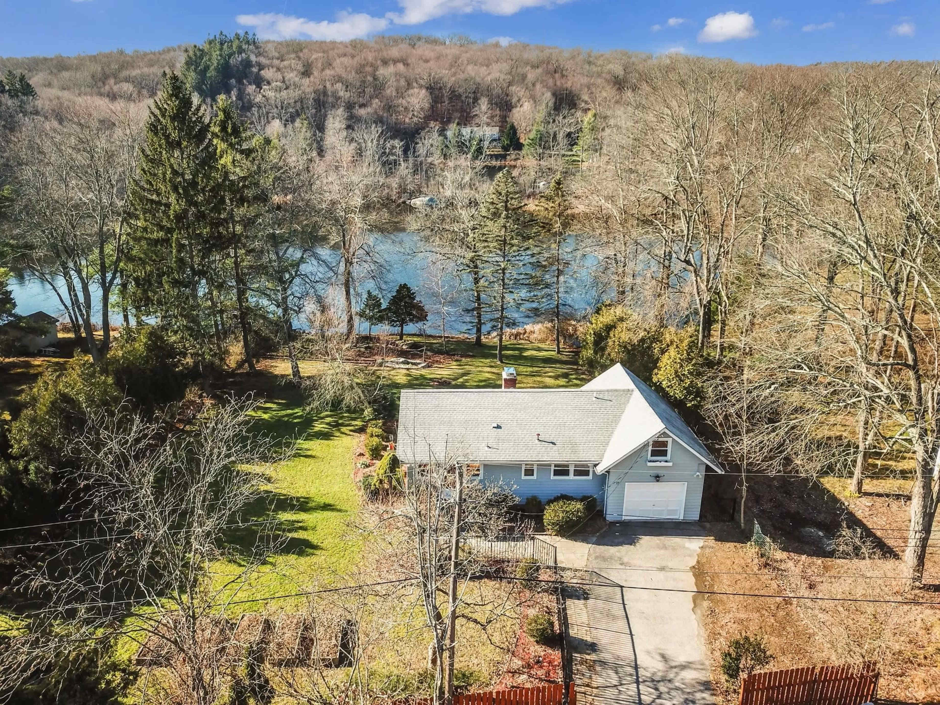 A single-story blue house with a garage is situated on a spacious lot near a calm body of water, surrounded by bare trees and rolling hills in the background.