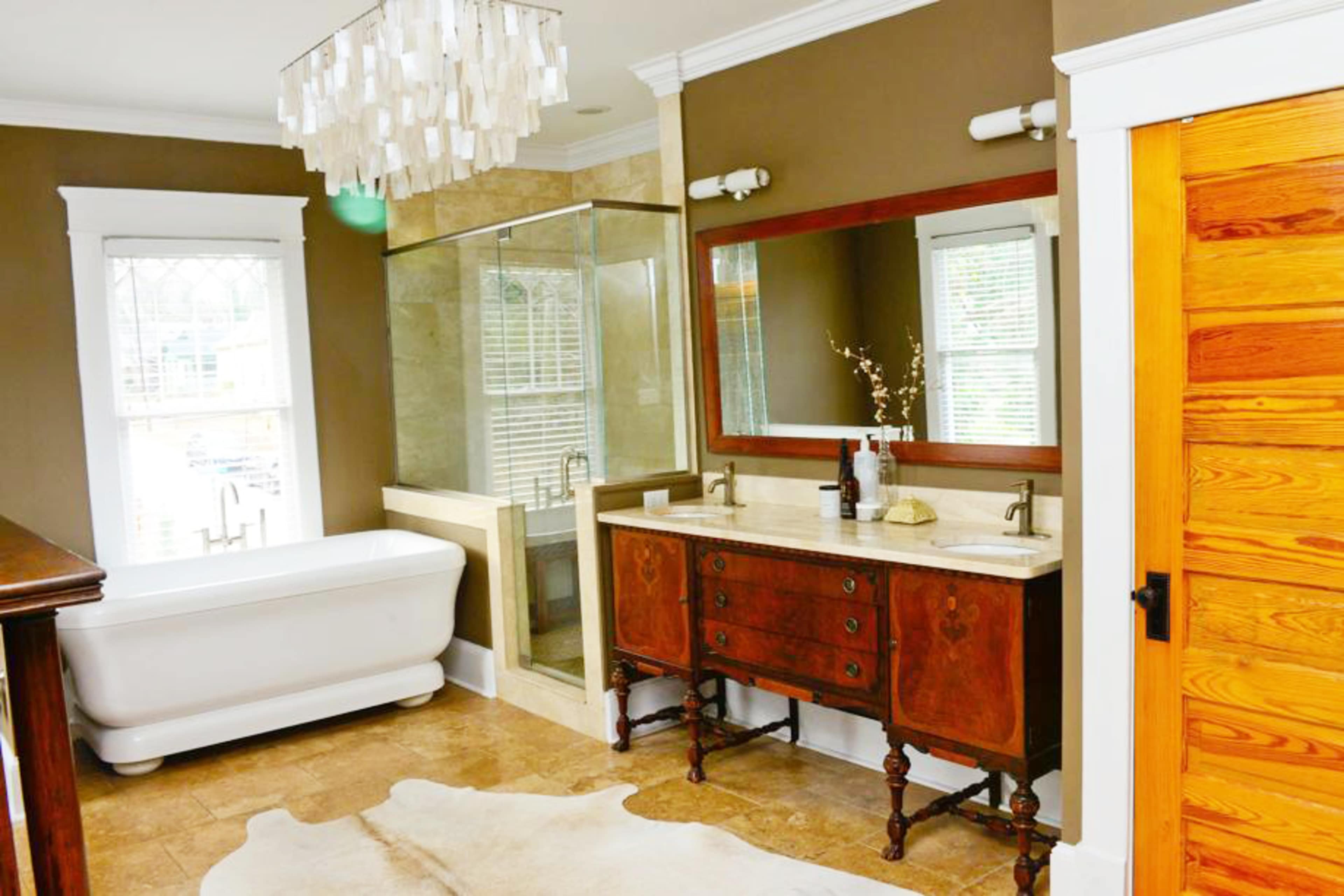 A modern bathroom features a freestanding bathtub, a walk-in shower with glass doors, a double vanity with a marble countertop, and natural light streaming in through a large window.