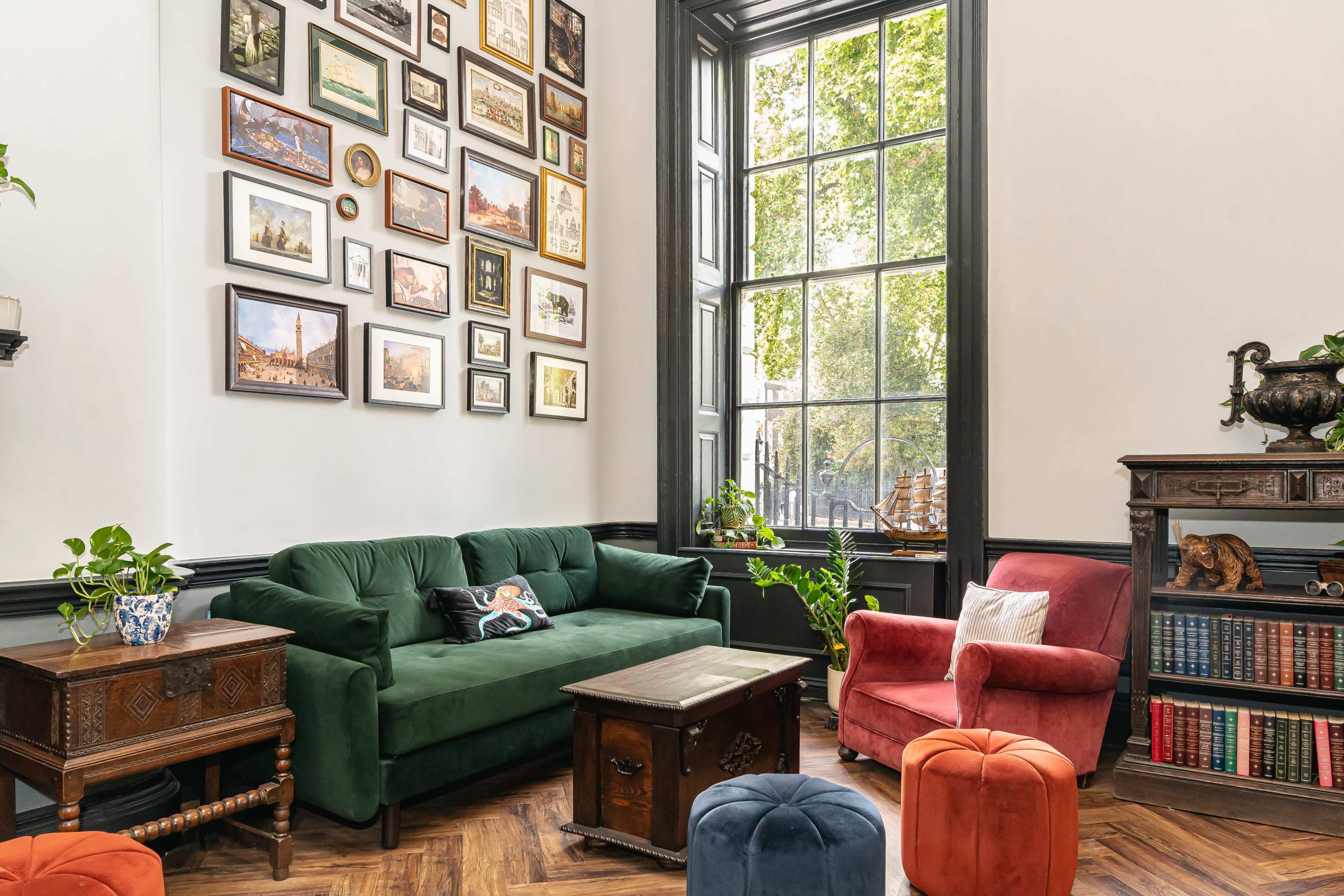 The image shows a stylish living room with a green sofa, red armchair, a wooden coffee table, and a gallery wall of framed pictures, illuminated by natural light from a large window.