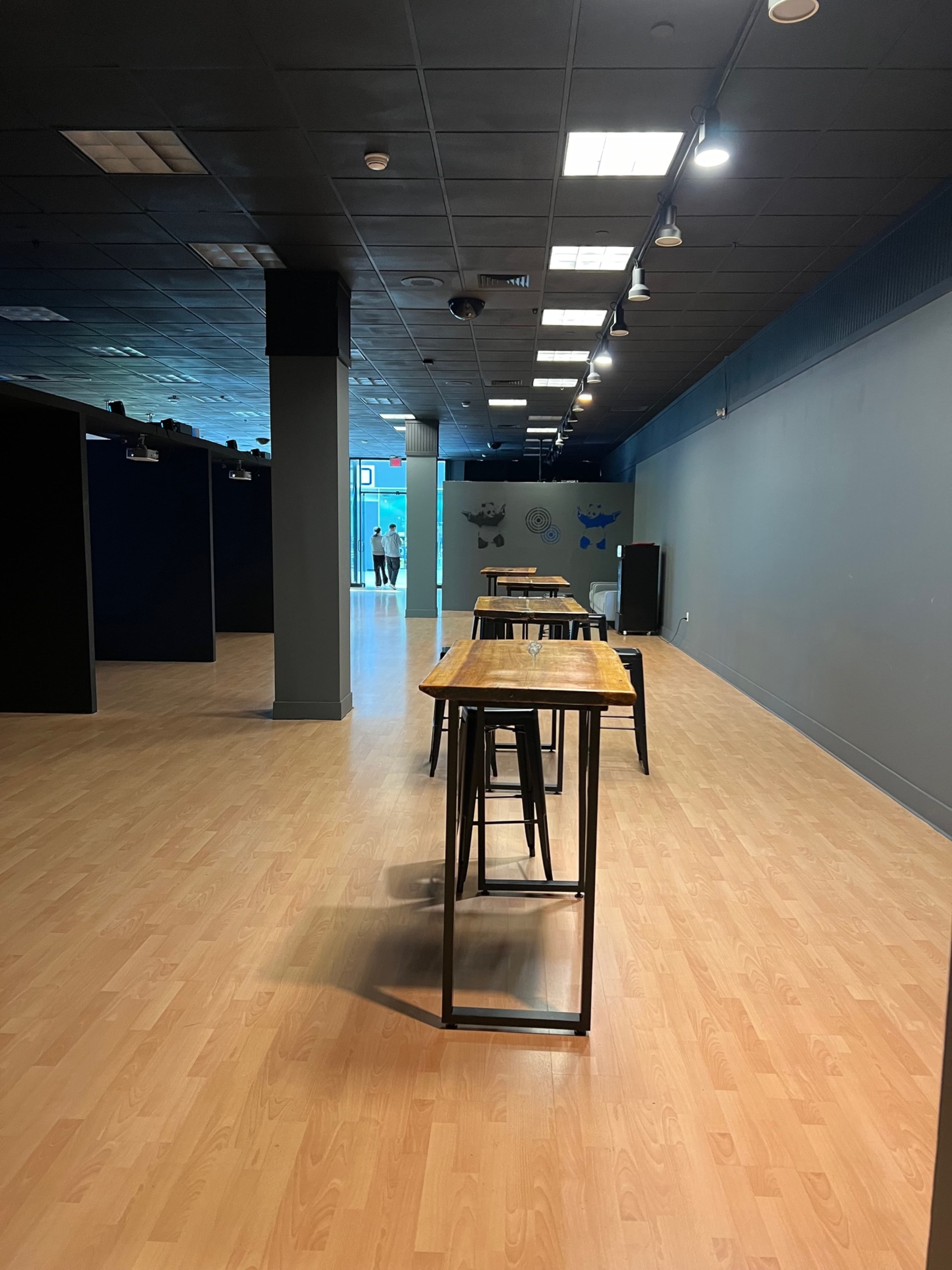 The image shows a spacious interior with wooden tables and stools arranged along a light-colored floor, leading to a door at the far end.