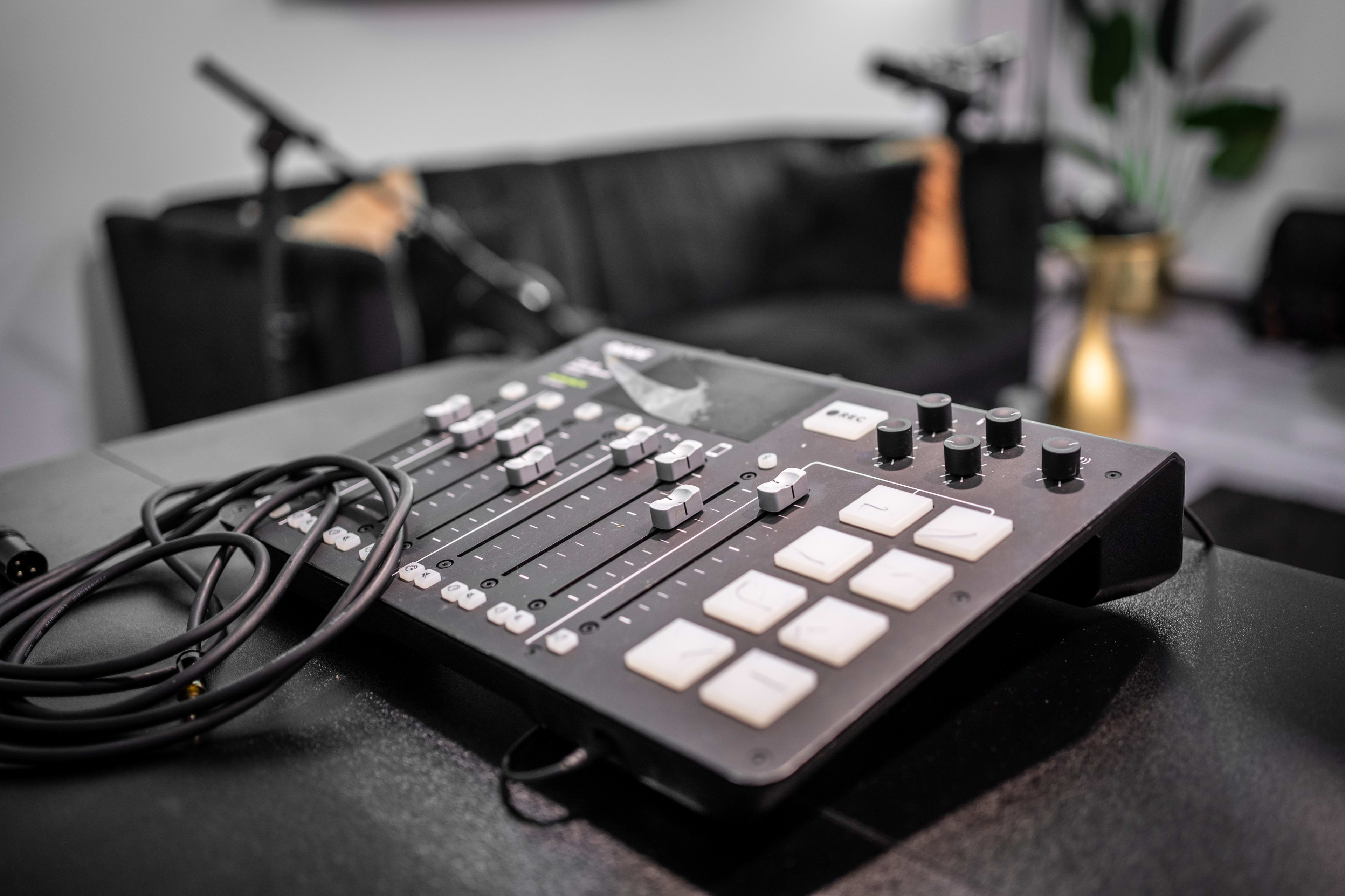 A close-up view of a sound mixing console with sliders and buttons, positioned in a dimly lit room with a couch and microphones in the background.