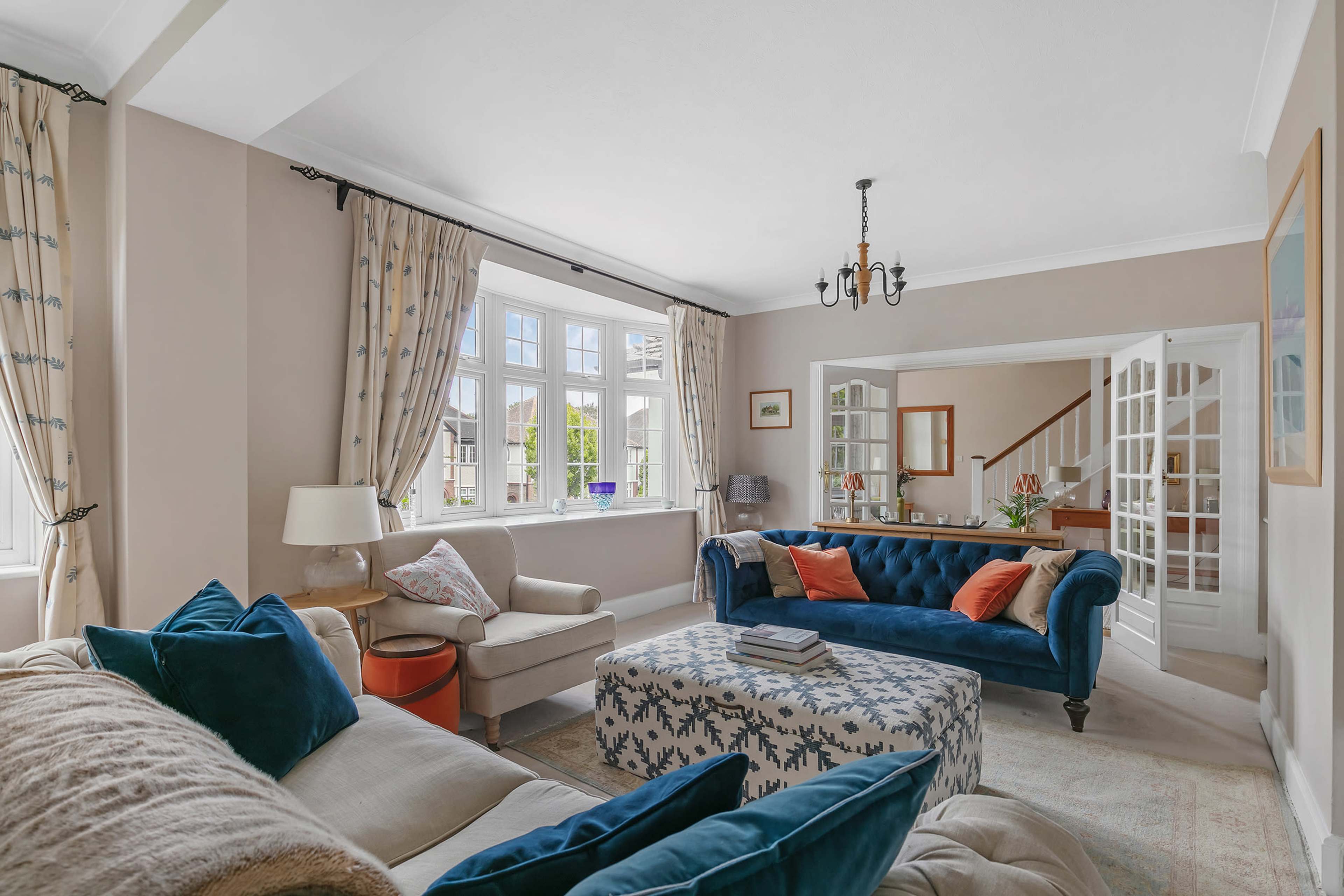 A spacious living room features a blue upholstered sofa, a patterned ottoman, and large windows dressed with curtains, allowing natural light to fill the space.