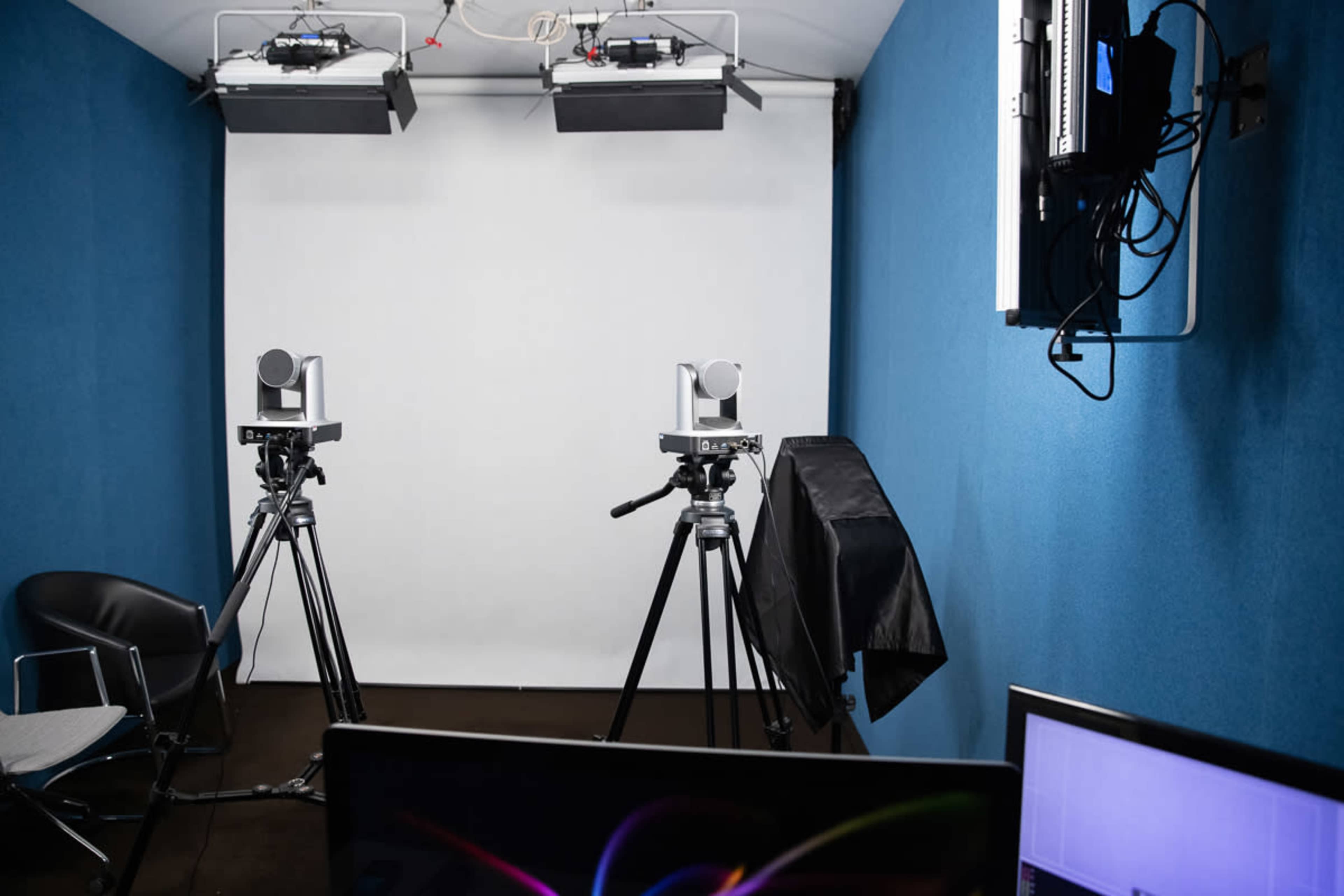 A small studio setup with two cameras on tripods facing a plain white backdrop, and a computer monitor visible in the foreground.