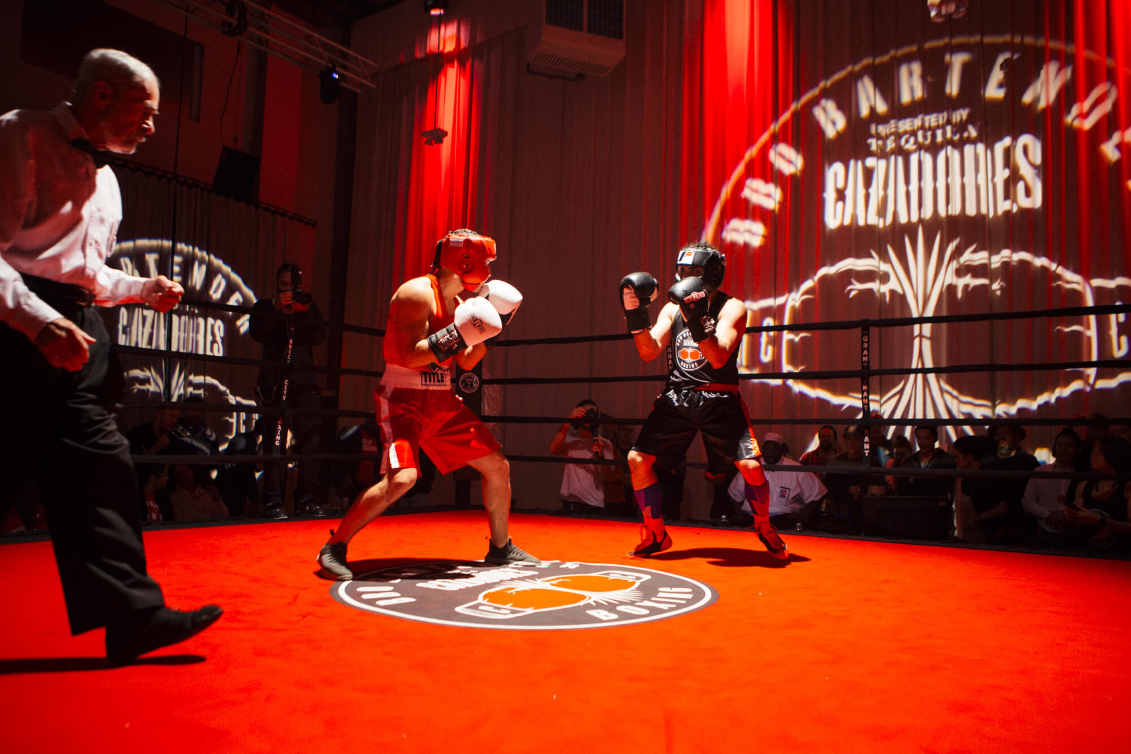 Two boxers are engaged in a match inside a well-lit ring, with spectators and an official present in the background.