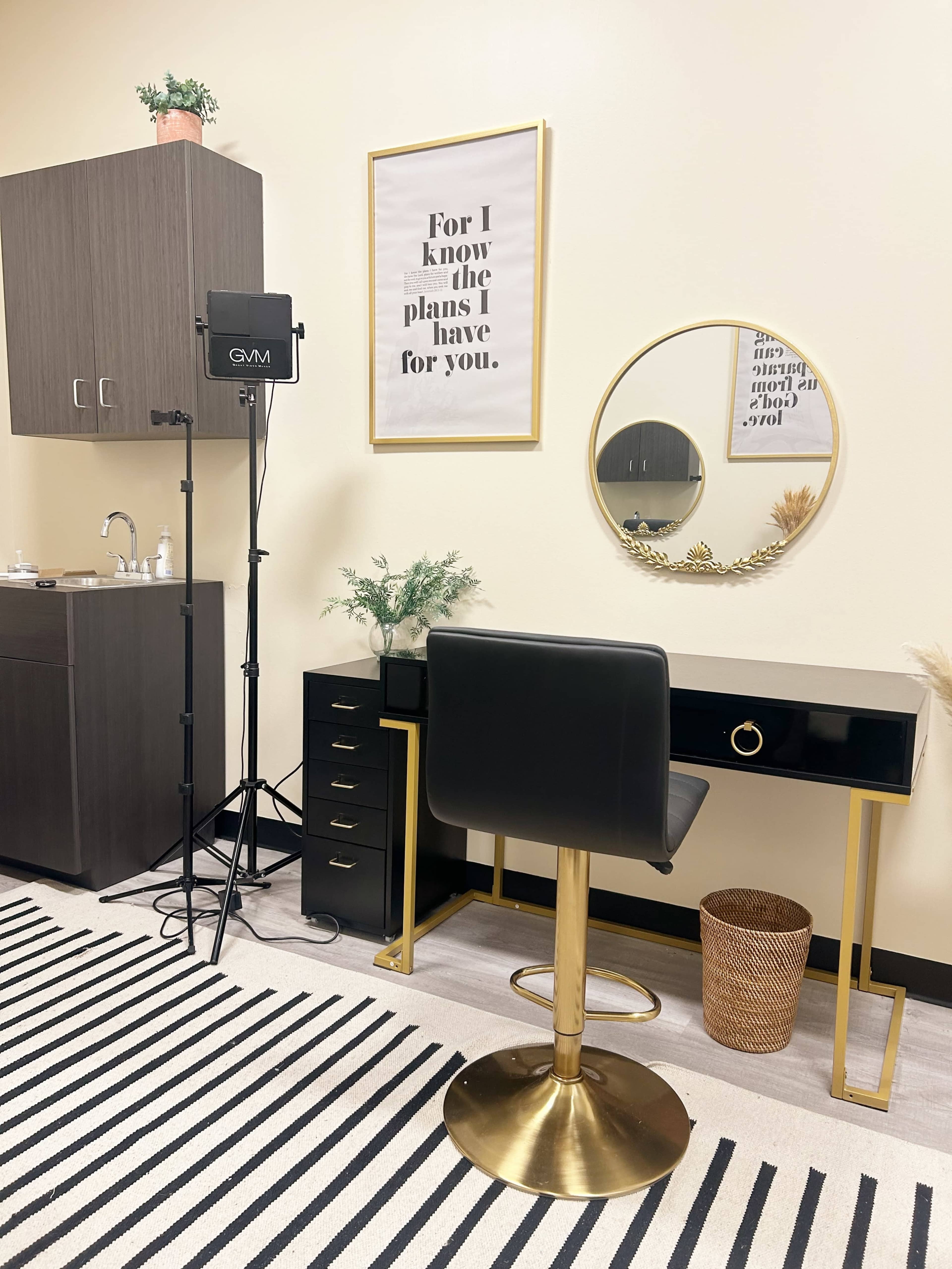 A modern workspace featuring a black and gold desk with a stool, a large round mirror, and a framed quote on the wall, alongside a plant and a standing light.
