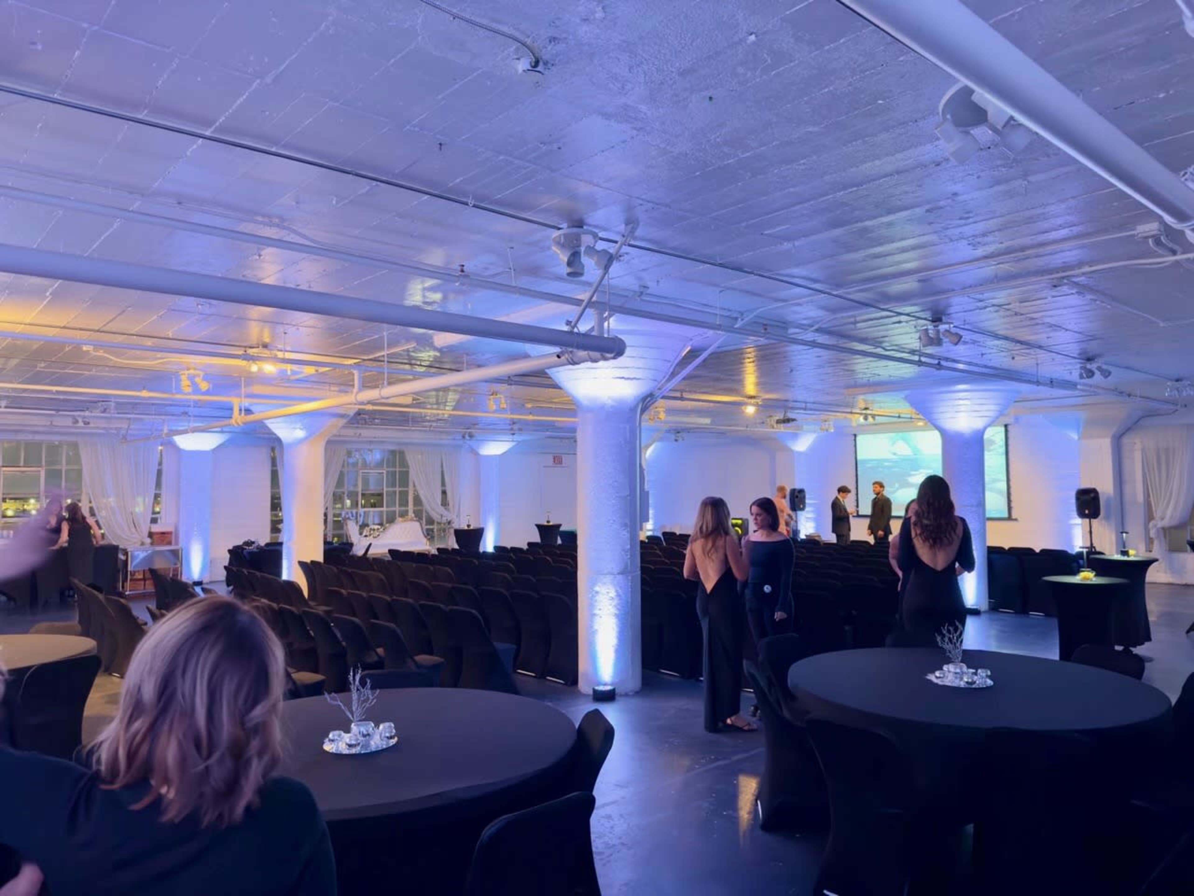 A spacious event venue is set up with black tables and chairs, illuminated by soft blue and white lighting, as attendees mingle in groups.