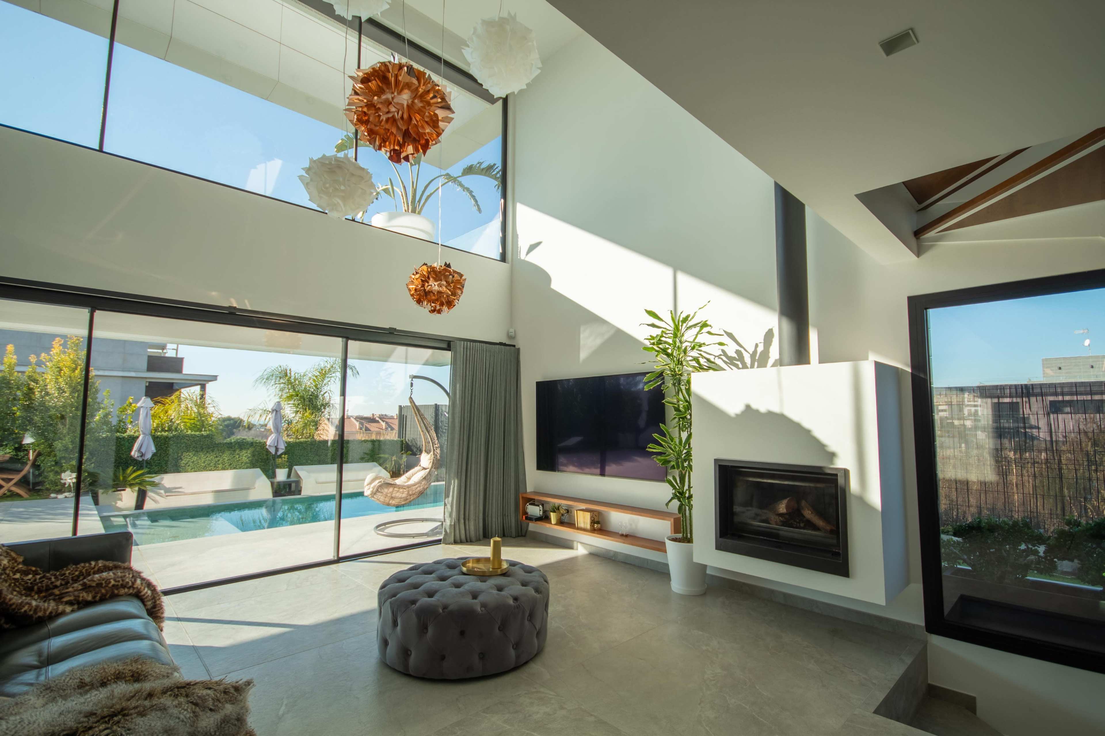 A modern living room with large windows overlooking a pool area, featuring a fireplace, a sectional sofa, and a round ottoman.