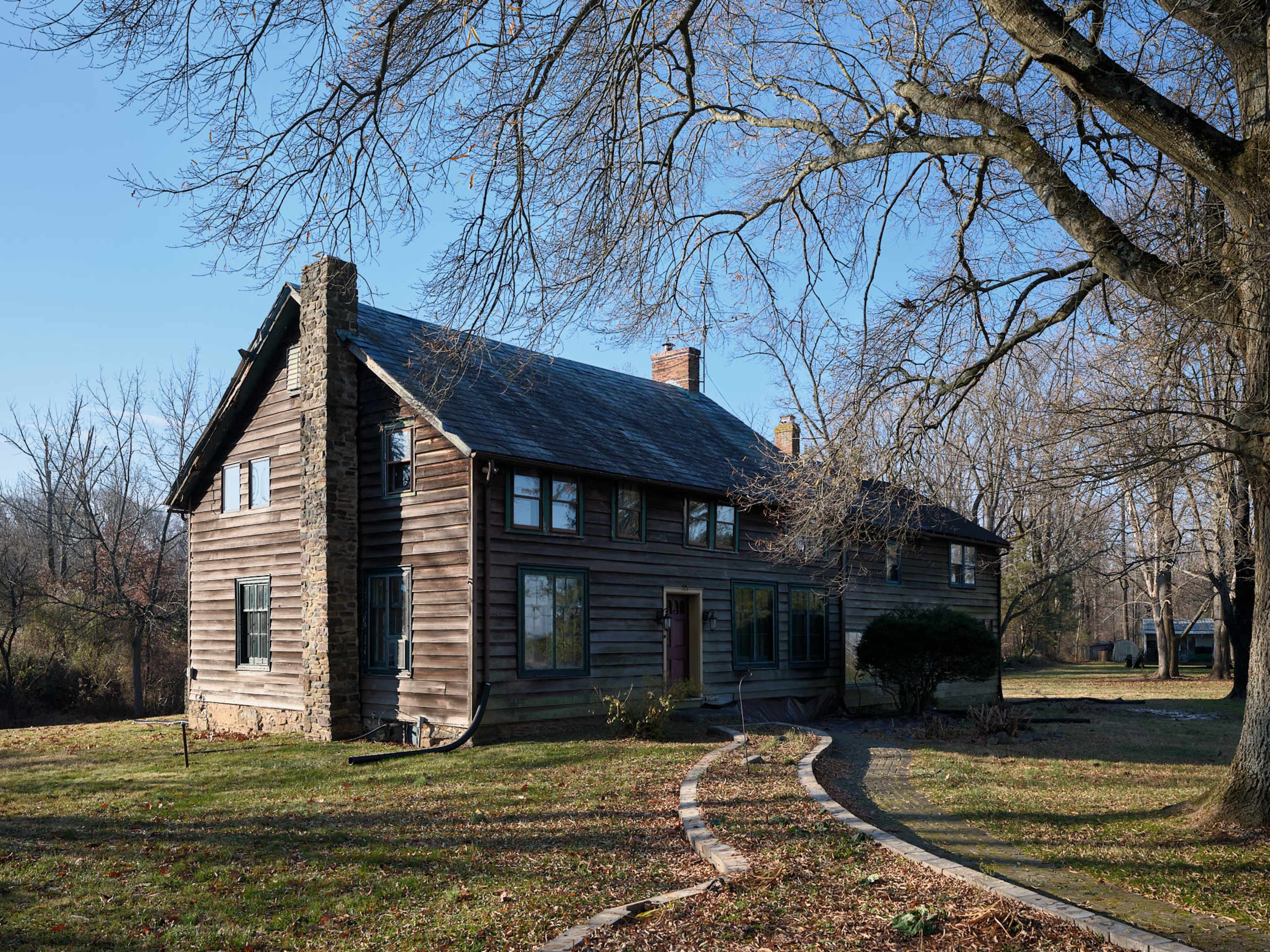 A two-story, wooden house with a stone chimney, set on a grassy plot with a winding stone pathway and bare trees in the background.