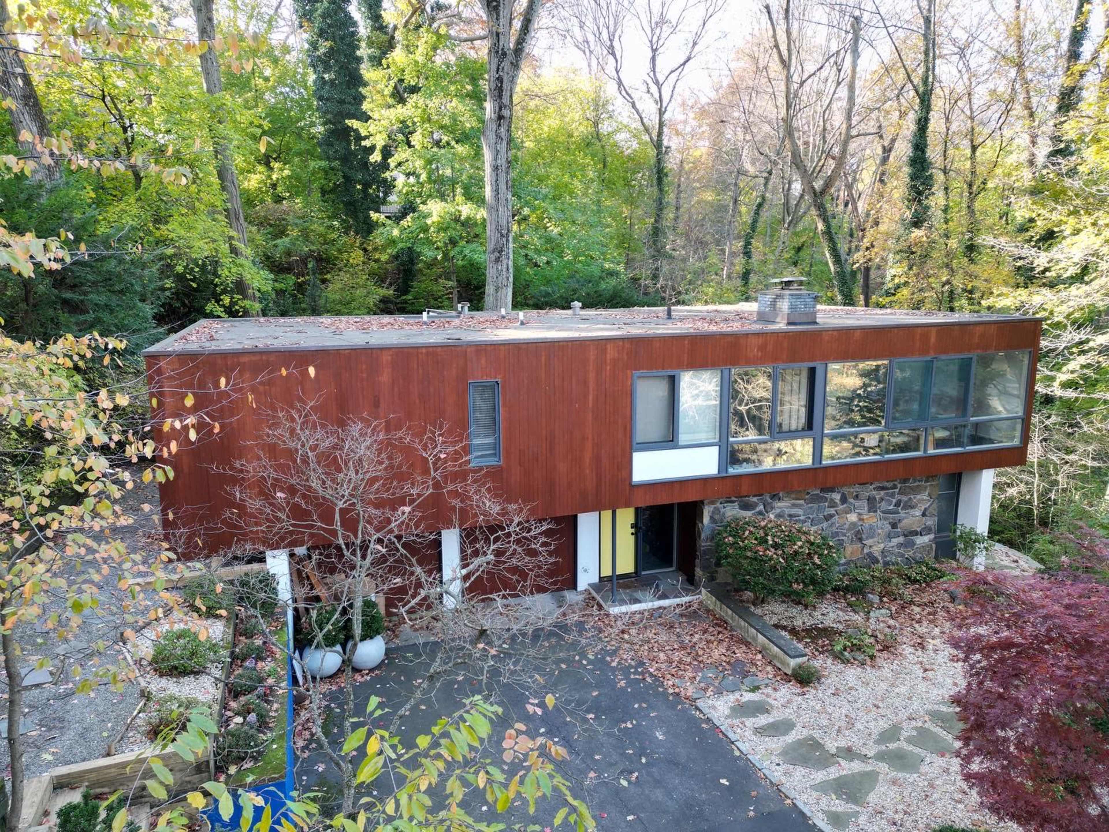 A modern two-story house with a wooden exterior and large glass windows is surrounded by trees in a wooded area.