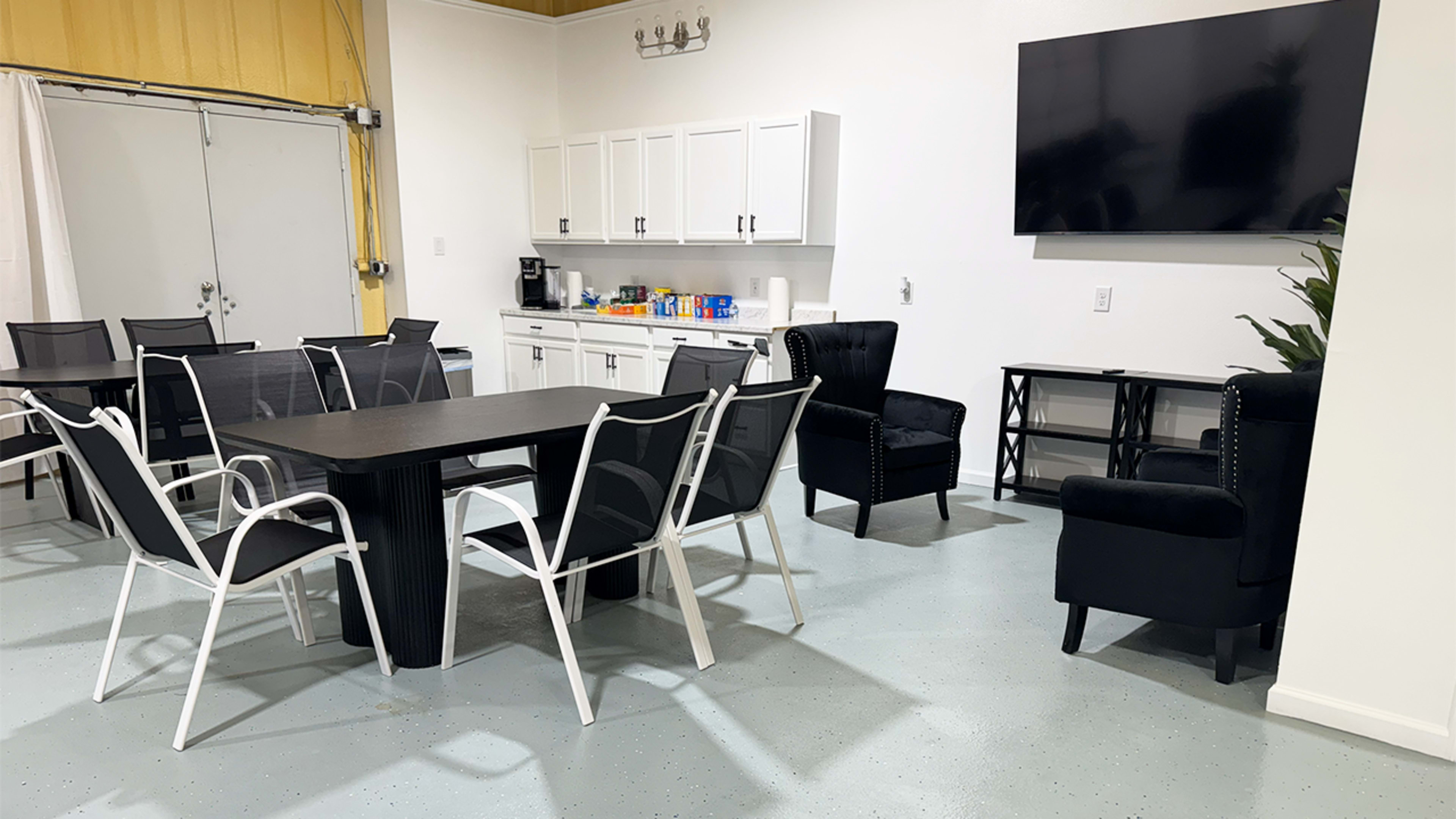 The image shows a modern breakroom with a large table, several chairs, a kitchenette area, and a wall-mounted television.